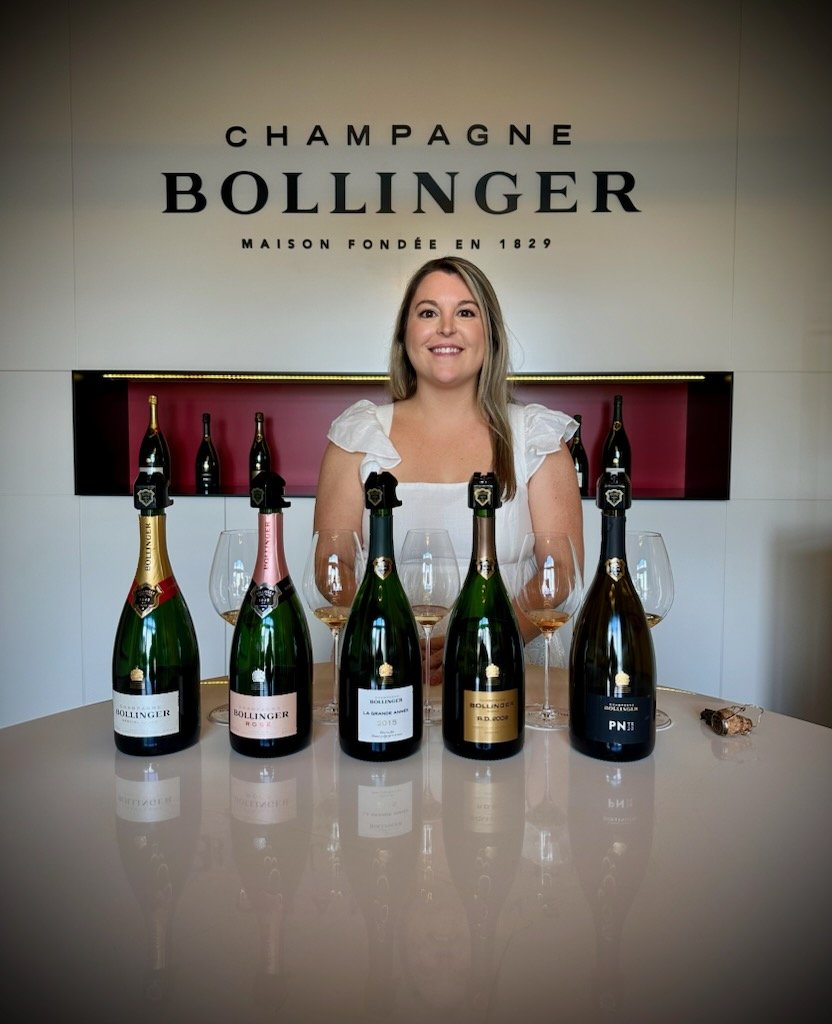 Woman smiling behind a table with five bottles of Bollinger champagne and five wine glasses at a champagne tasting event.