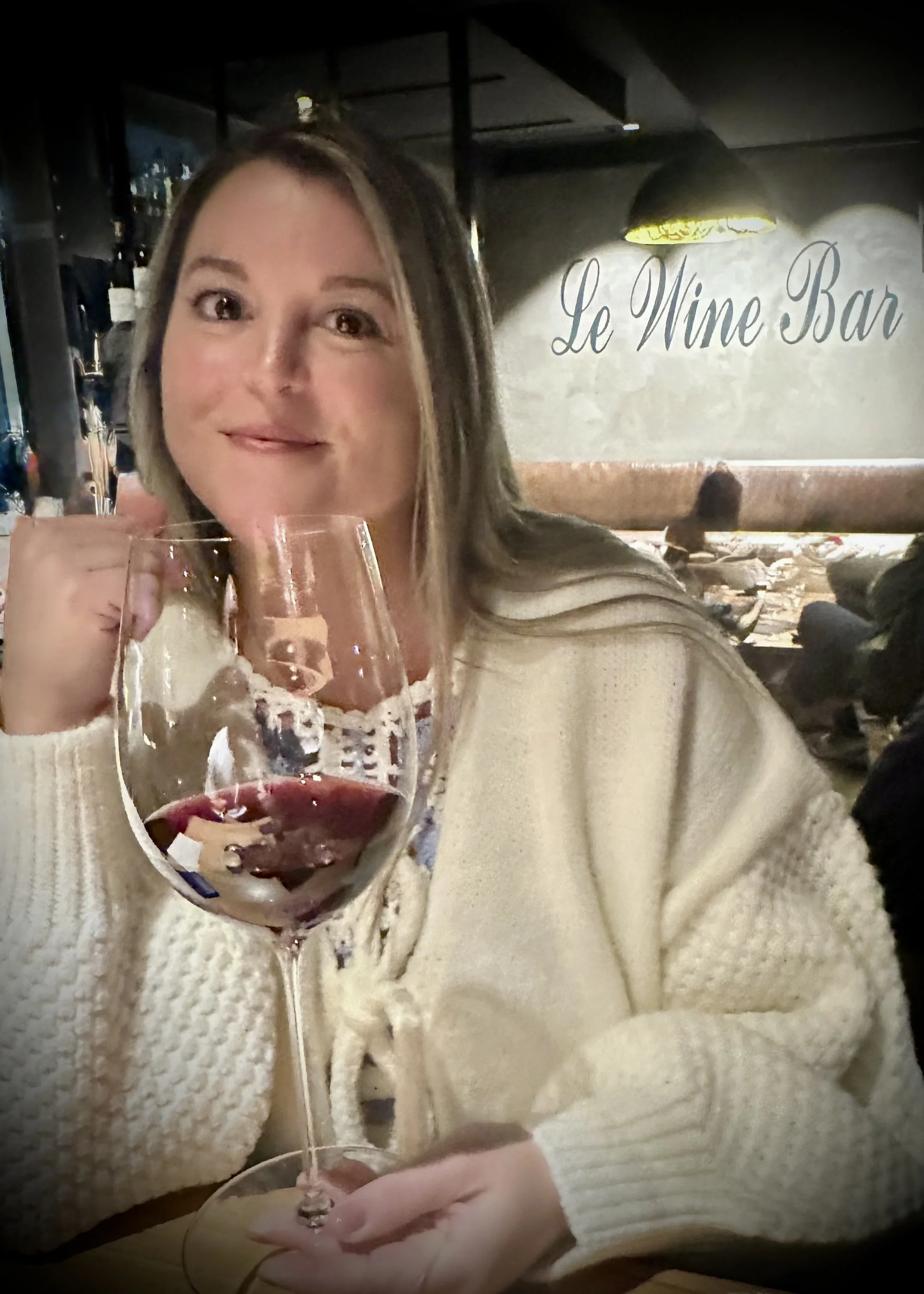A woman with shoulder-length hair holding a glass of red wine at Le Wine Bar, wearing a cream-colored sweater.