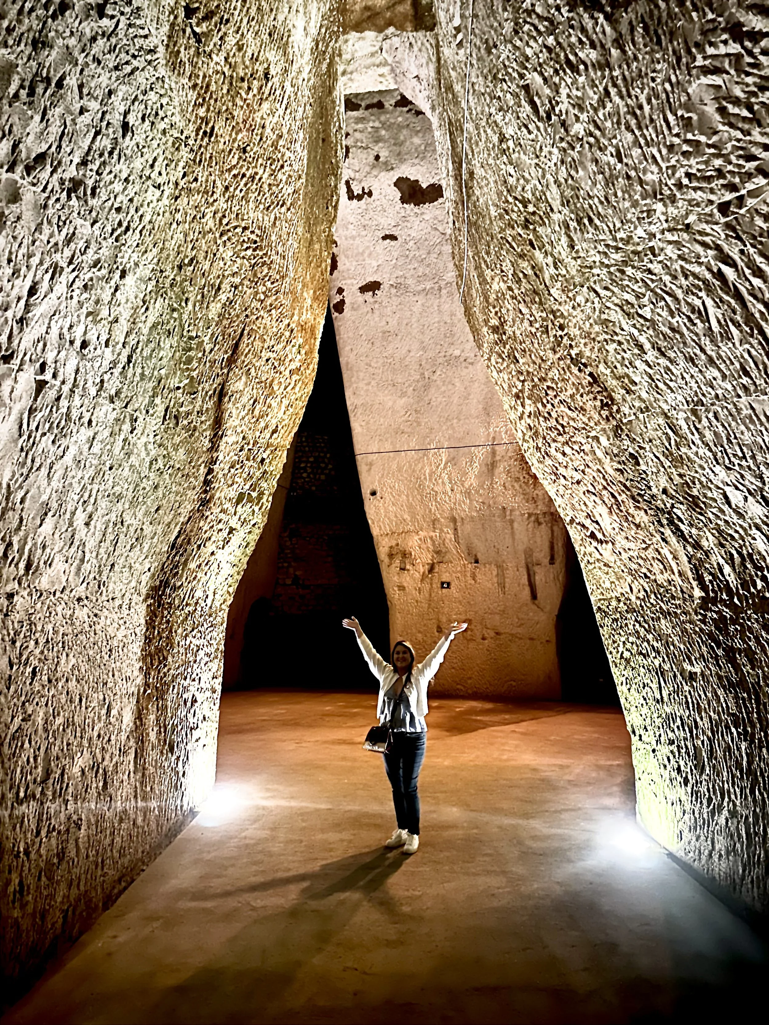A woman standing inside a large ancient stone structure with high, textured walls, smiling with arms raised.
