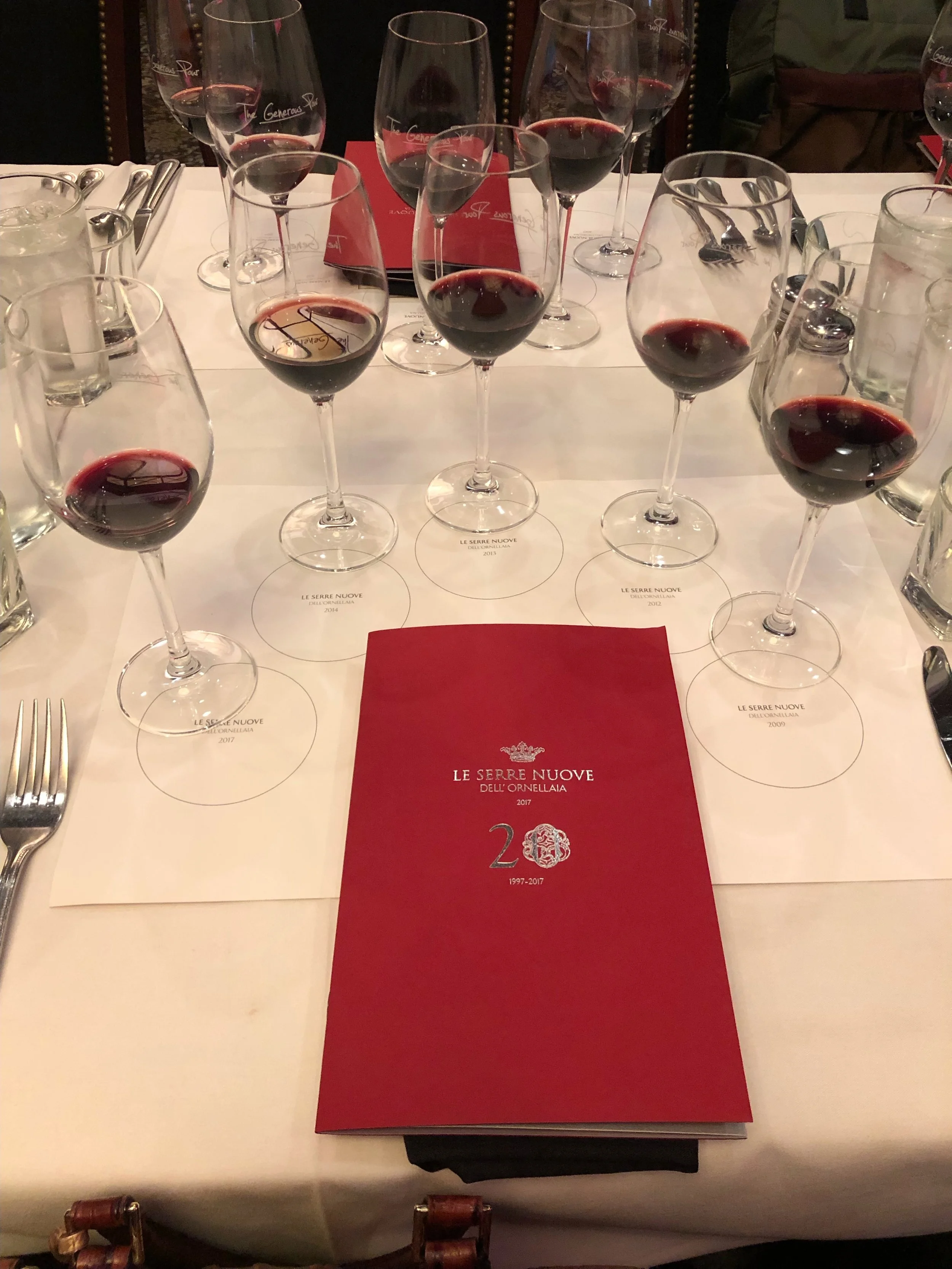A dining table with multiple glasses of red wine, a red menu, and some silverware in a restaurant setting.