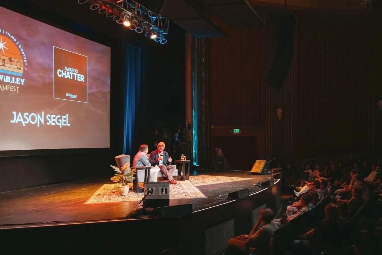 Scene from an awards event on a stage with two people seated in conversation, one with a microphone. The stage is decorated with a large screen displaying logos and text, including 'NAPA Valley STREAMFEST,' 'AWARDS CHATTER,' and 'JASON SEGL.' An audience is seated in front of the stage in a darkened theater.