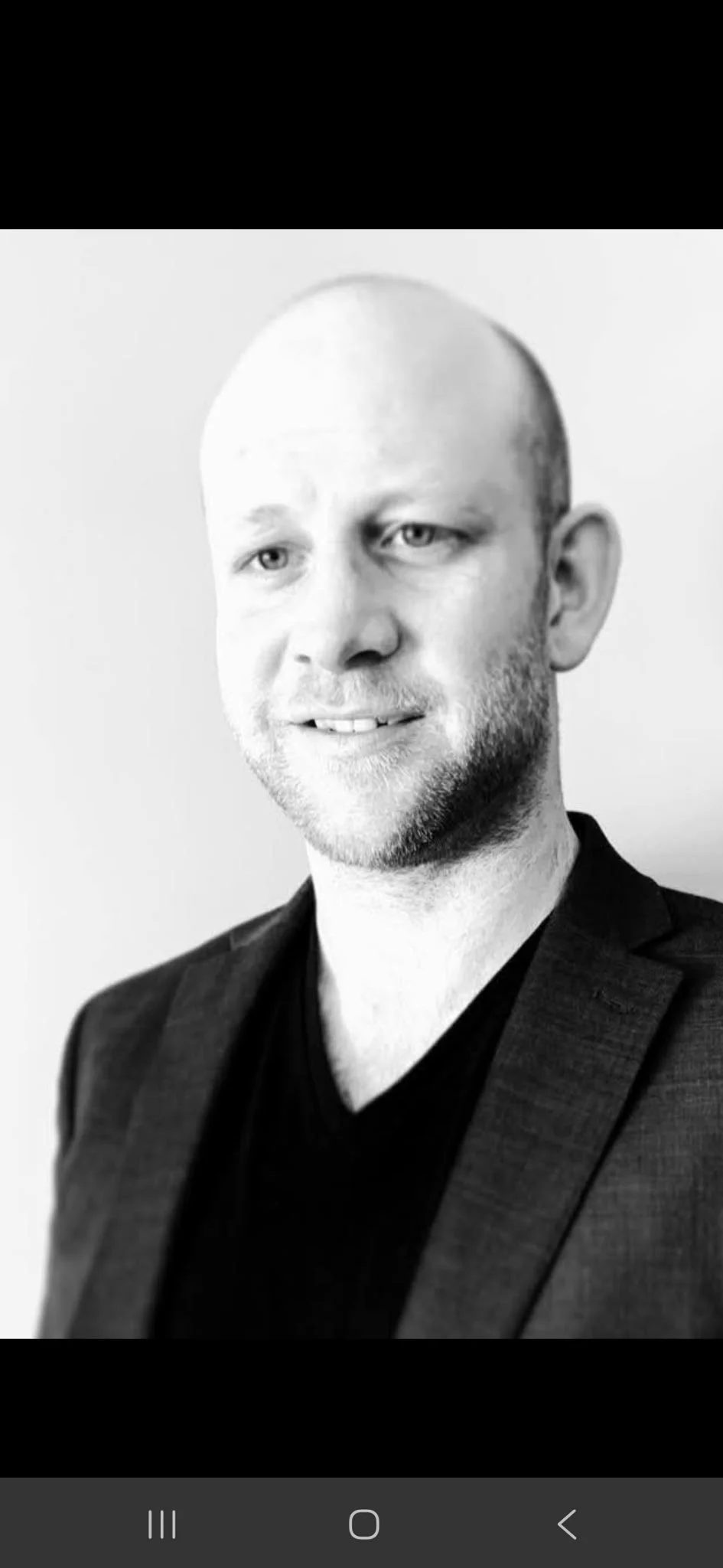 Black and white portrait of a man with a beard, wearing a dark blazer and a V-neck shirt, smiling slightly.