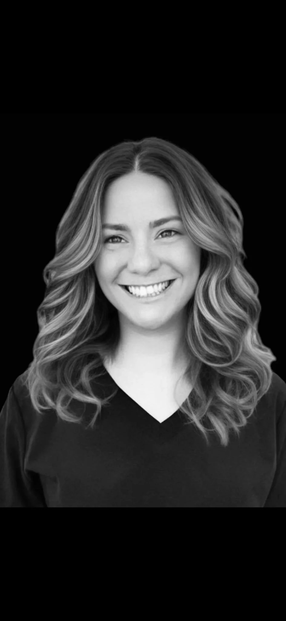 Black and white portrait of a smiling woman with wavy hair wearing a dark top, set against a black background.