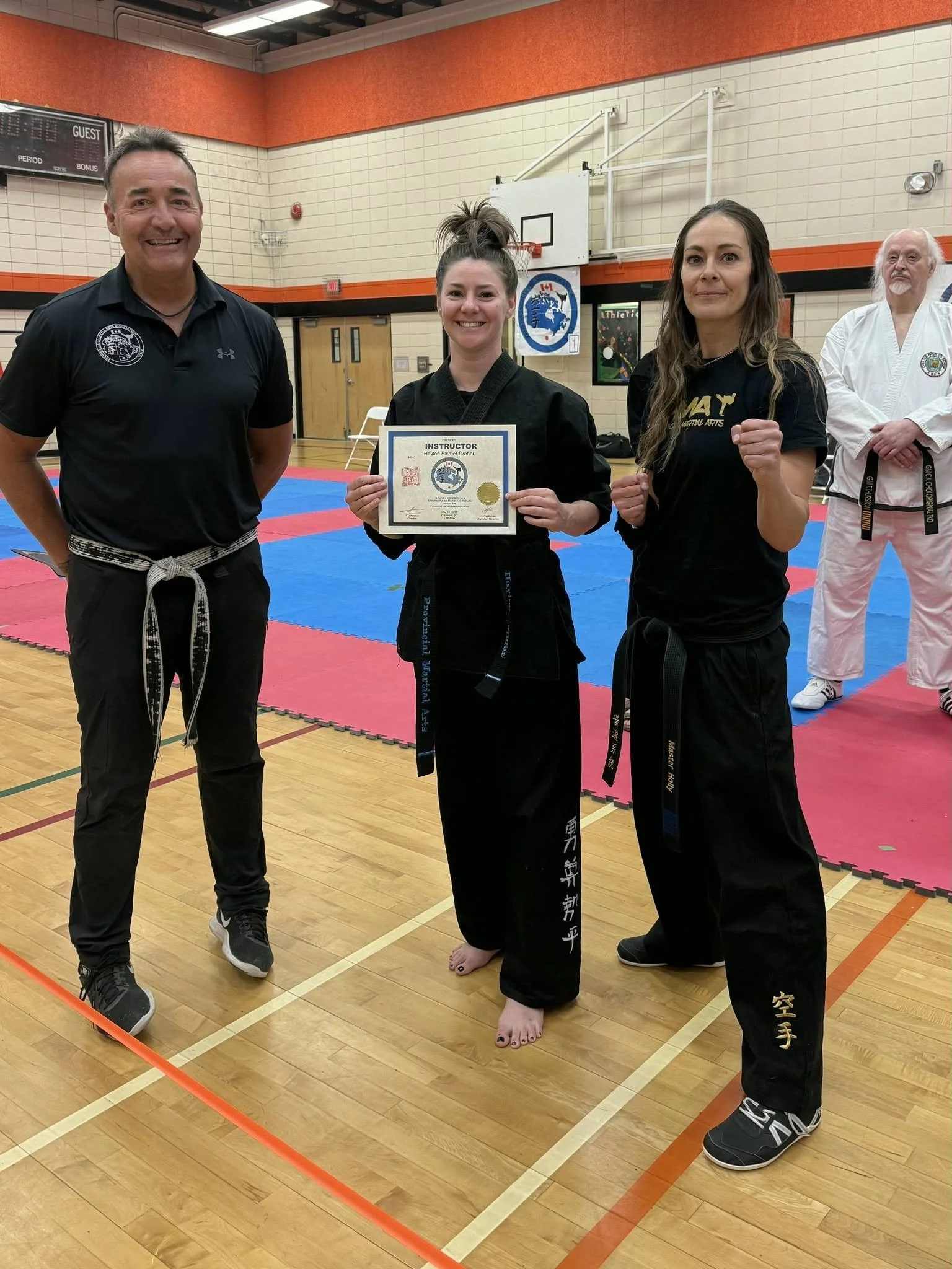 Group of martial arts practitioners standing on a gymnasium floor, with a young woman holding a certificate and a gold medal, flanked by two adults, one man and one woman, all dressed in martial arts uniforms.