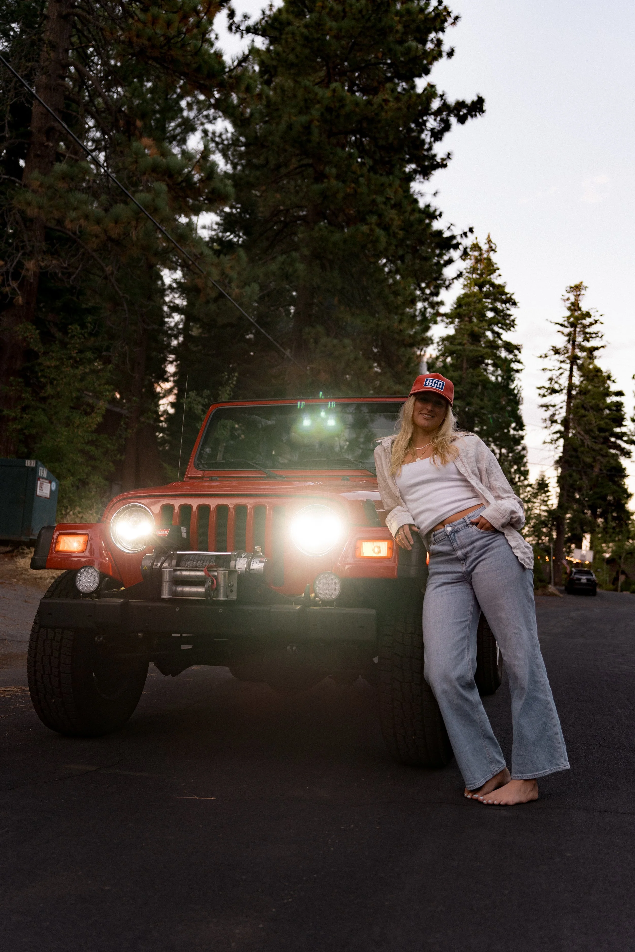 A woman in casual clothes, wearing a cap and denim jeans, leaning against a red Jeep parked on a driveway with trees in the background, during dusk.