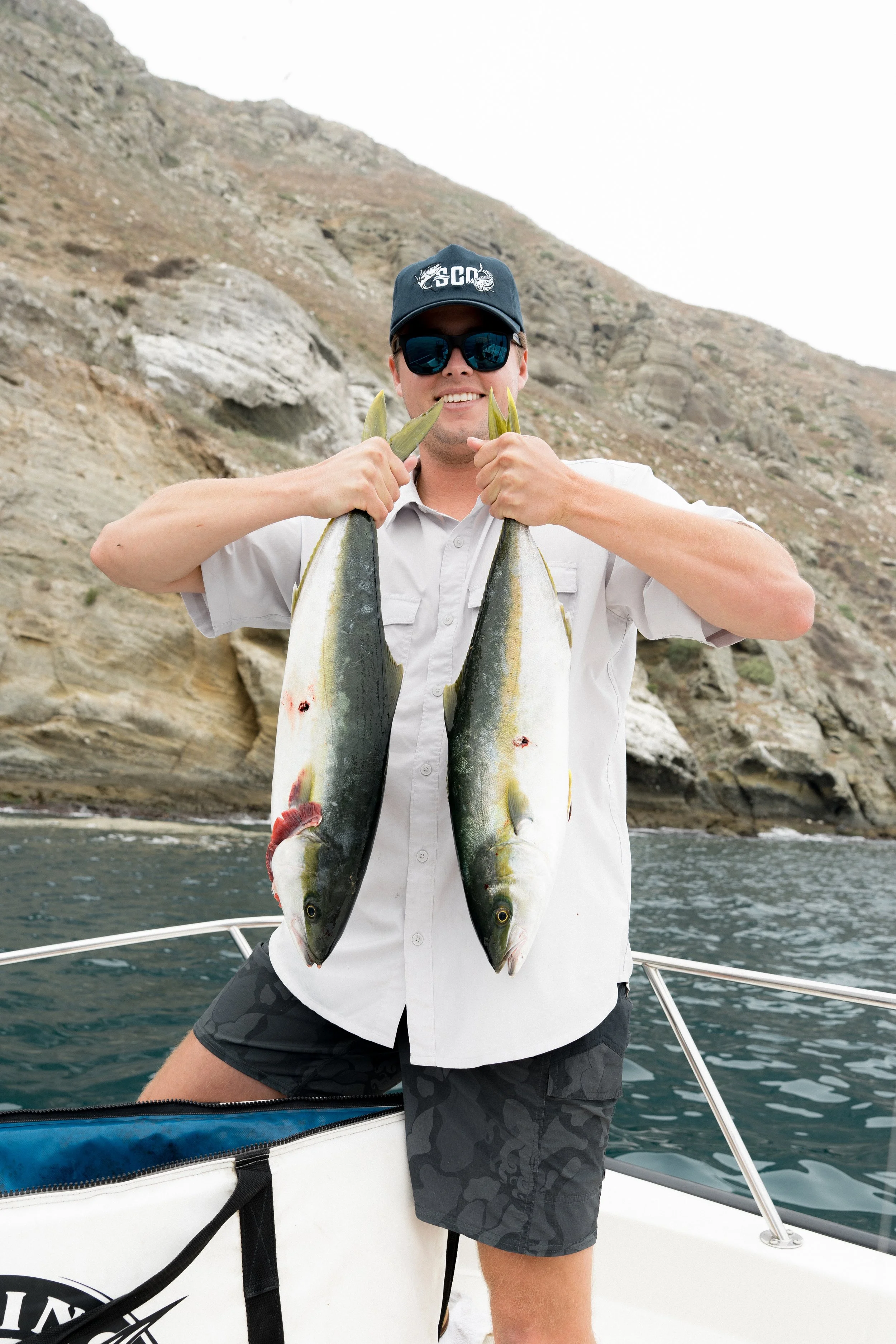 A man holding two large fish on a boat with rocky hills in the background.