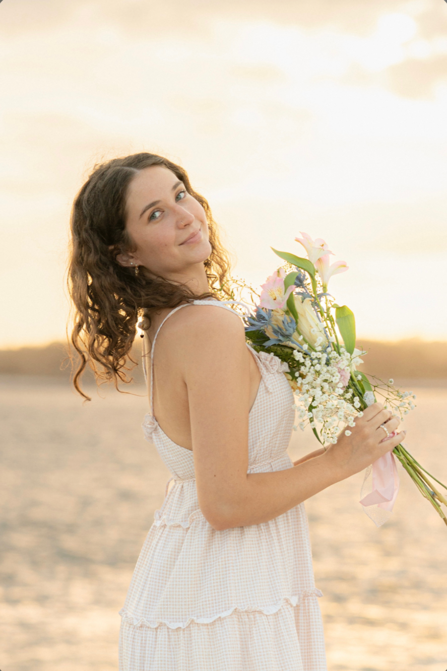 A young woman with shoulder-length brown hair smiling and looking  over her shoulder holding a bouquet of flowers.