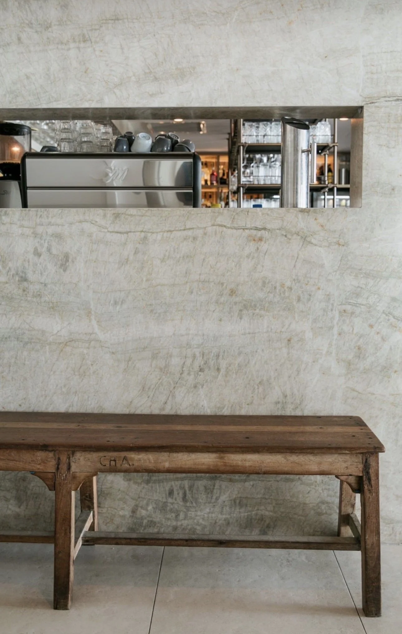 A wooden table in front of a textured stone wall, with a marble ledge and a metal shelf with glassware and bottles behind it.