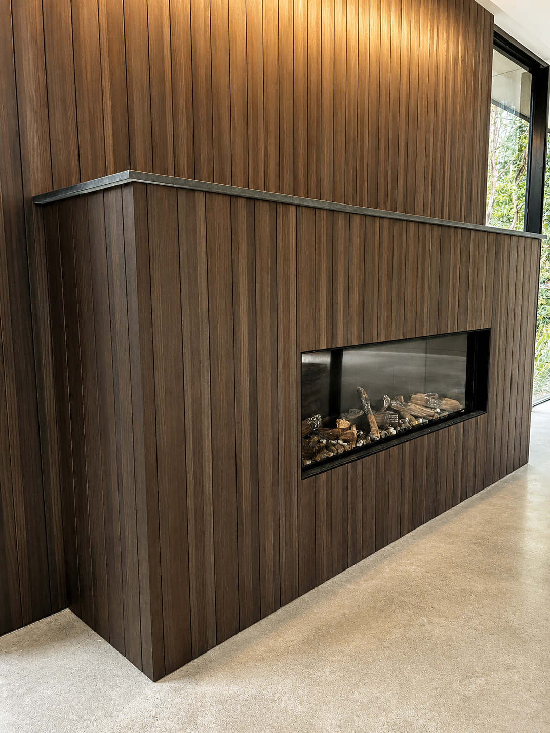 Wood-paneled interior wall with built-in rectangular fireplace filled with logs, next to large glass windows showing outside greenery.