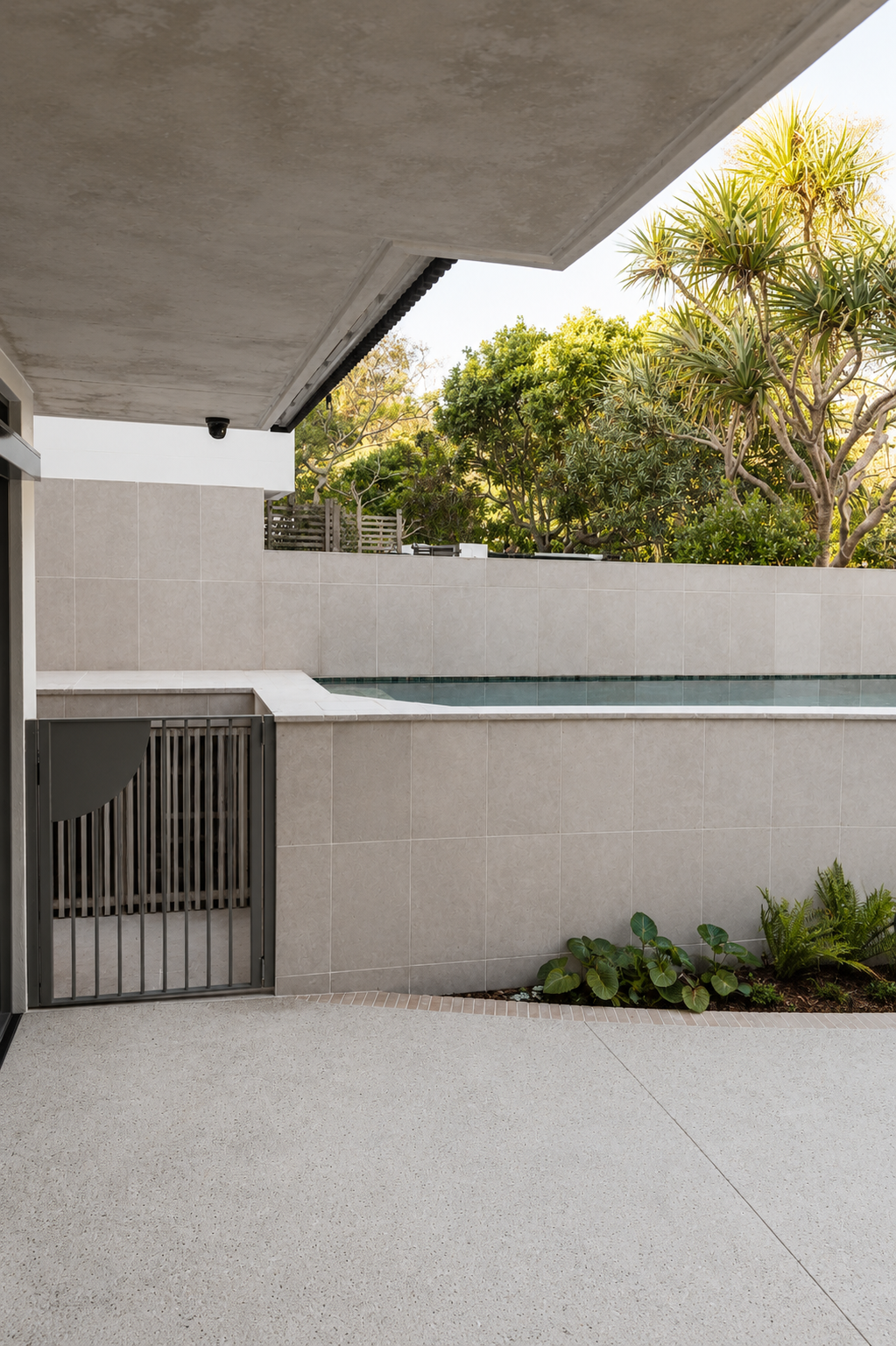 Modern balcony with a view of a pool, beige tiled wall, lush greenery, and trees in the background.