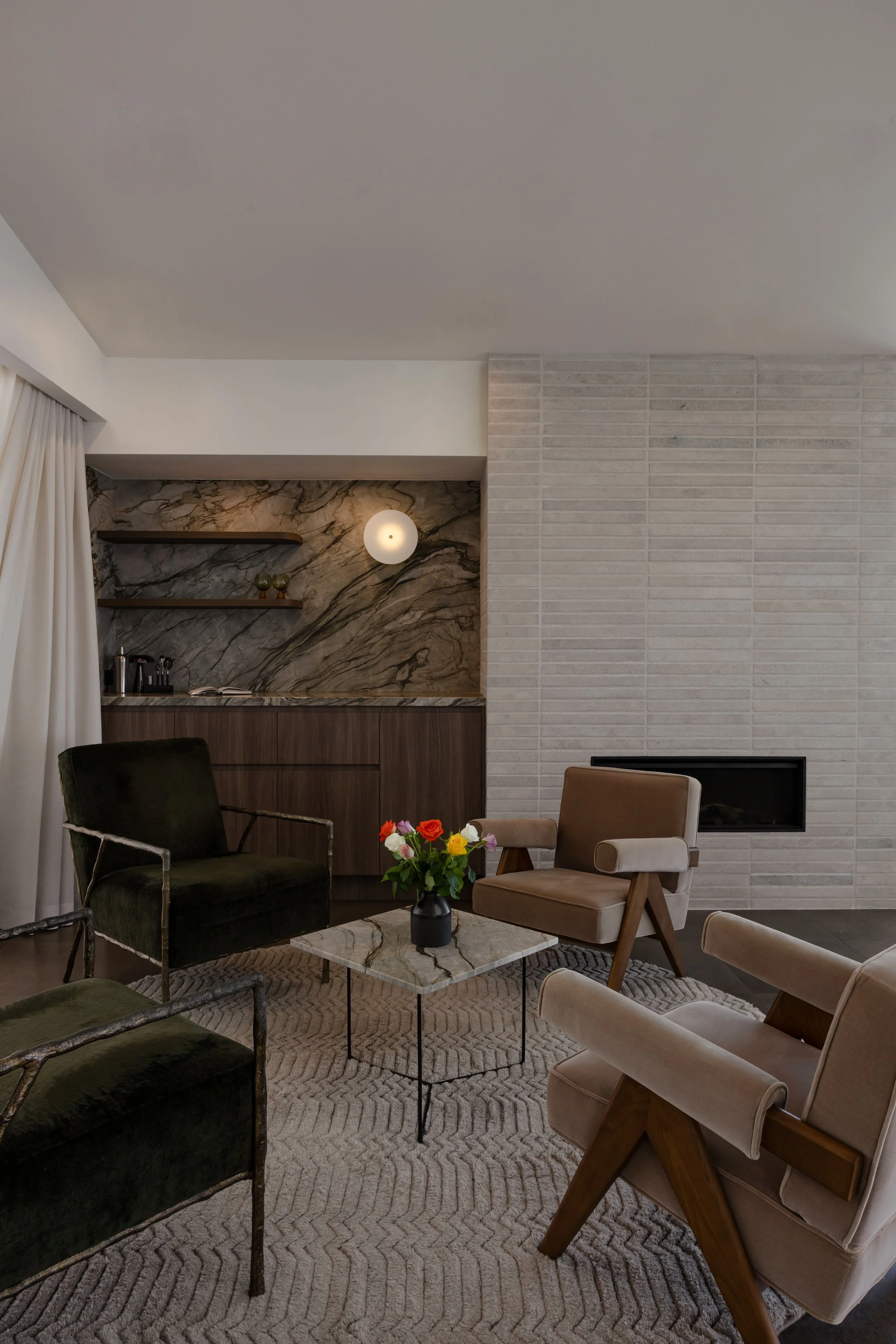 Living room with modern chairs, a marble coffee table with a flower vase, a textured rug, a built-in shelf with decorative items, and a wall with a combination of marble and brick textures.