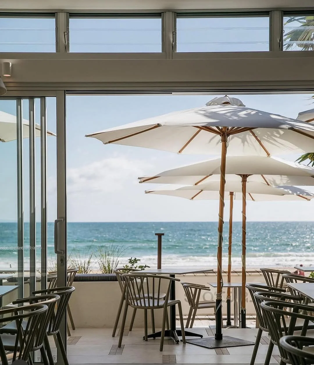 Indoor seating area with chairs and tables, large windows open to a beach view with umbrellas, the ocean, and a partly cloudy sky.