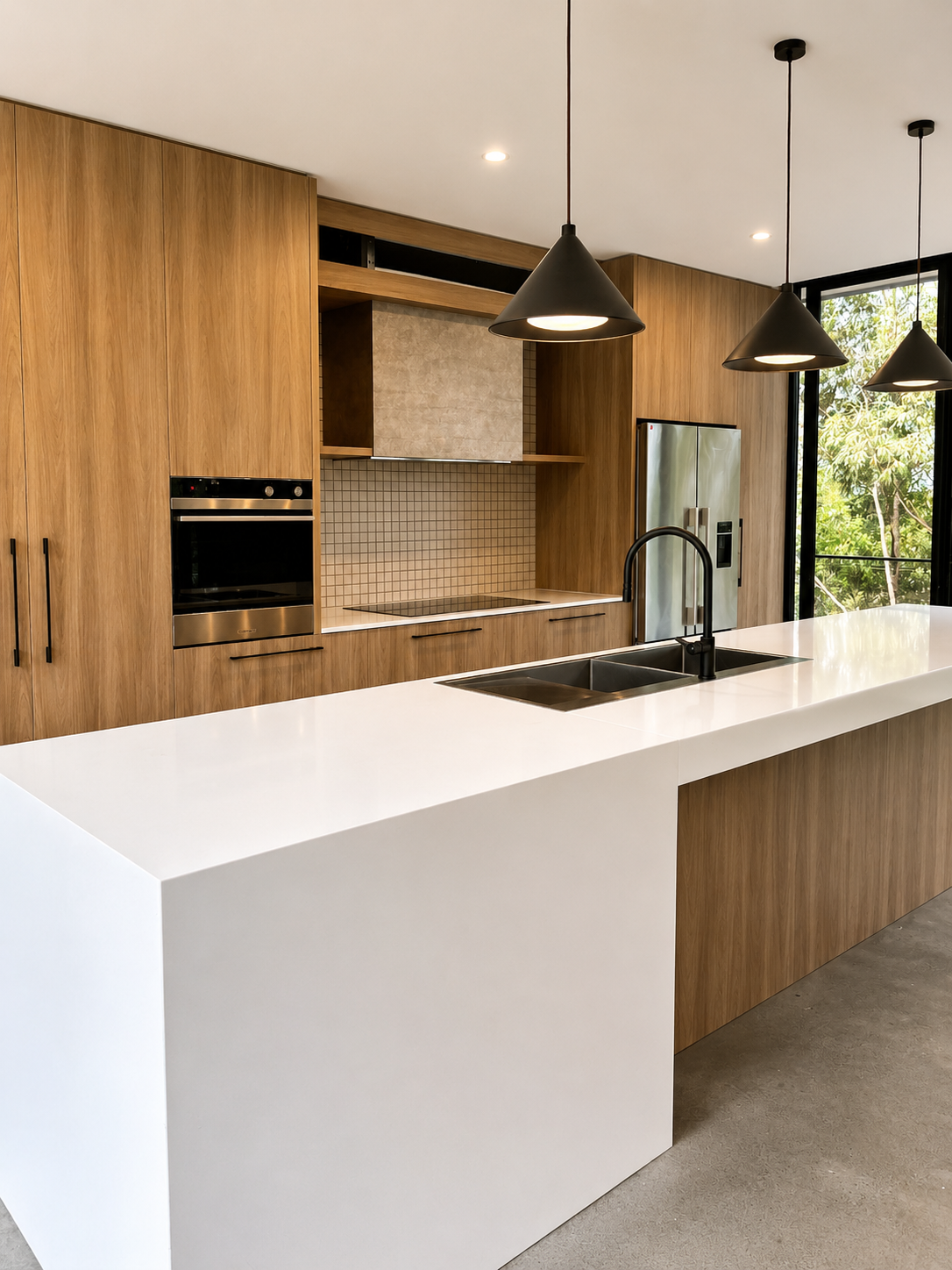 Modern kitchen with wooden cabinets, white island counter, black pendant lights, stainless steel appliances, and large window showing greenery outside.