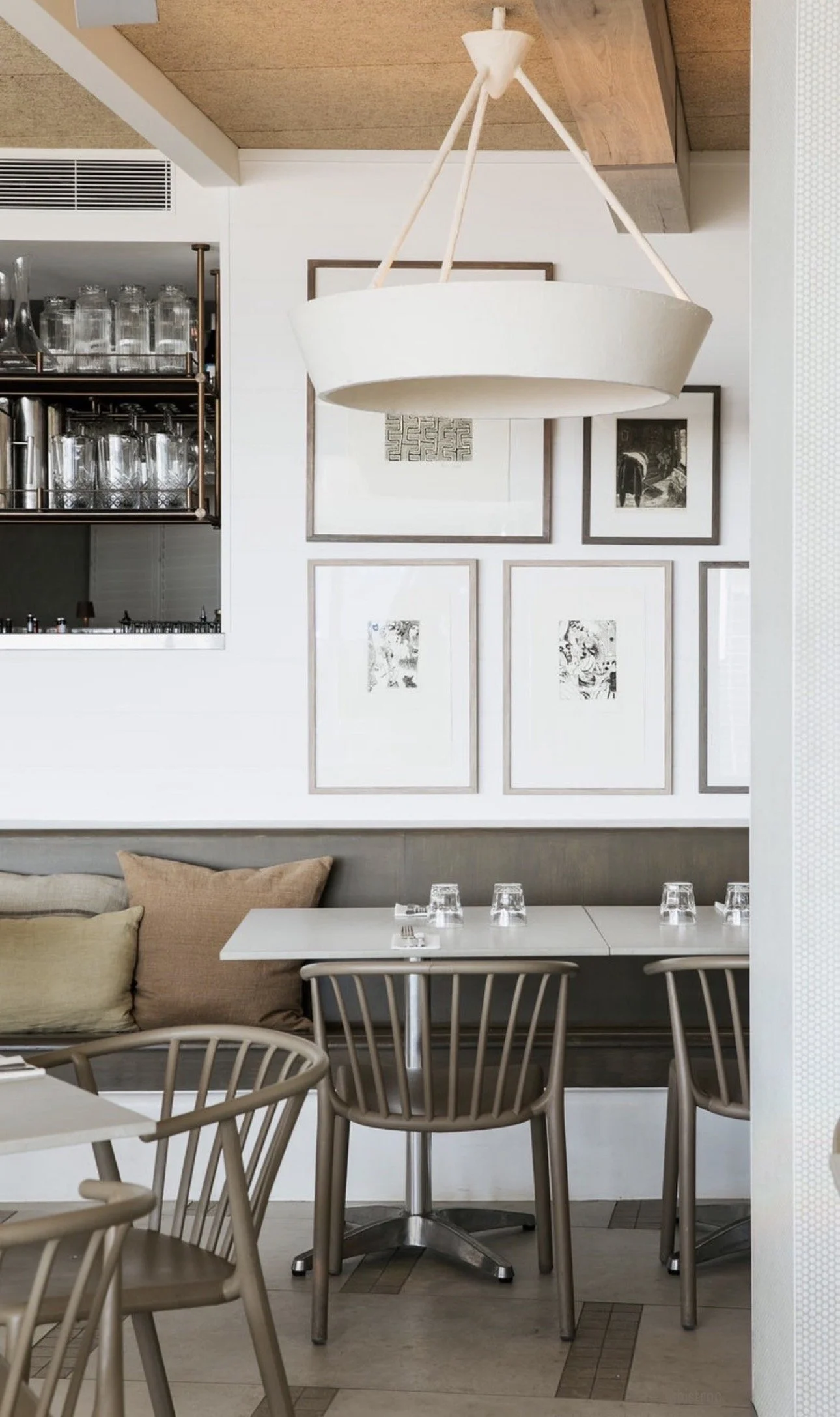 Interior of a modern restaurant with minimalist decor, featuring a white hanging light fixture, framed black and white artwork on the wall, and tables set with glasses and cutlery.