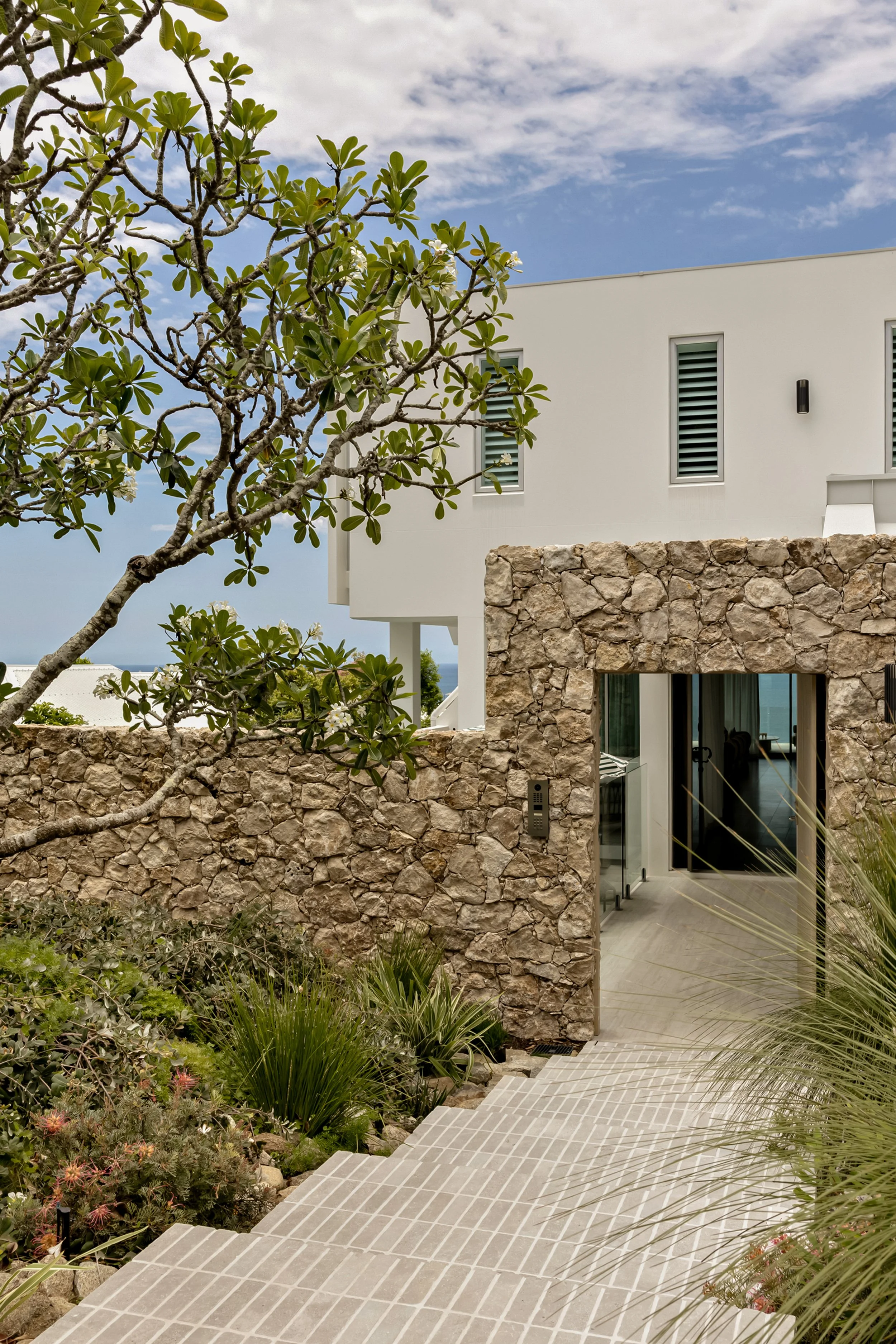 Exterior view of a modern white house with large windows and a stone wall, surrounded by lush green plants and trees, with steps leading to a patio area, under a partly cloudy sky.