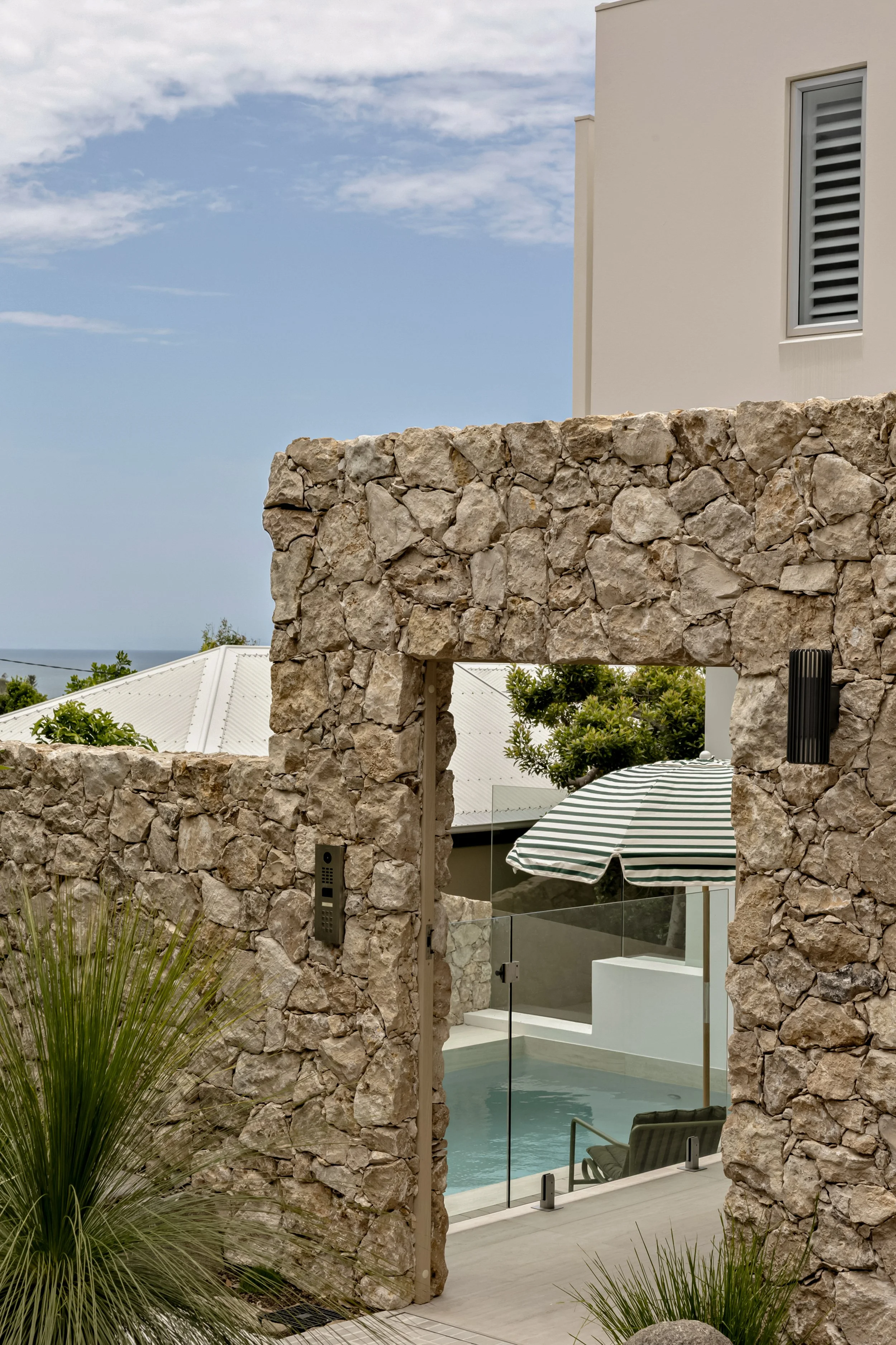 View of a modern house with a stone archway in the foreground, overlooking a swimming pool area with a striped patio umbrella, cooling chairs, and lush greenery, under a partly cloudy sky.