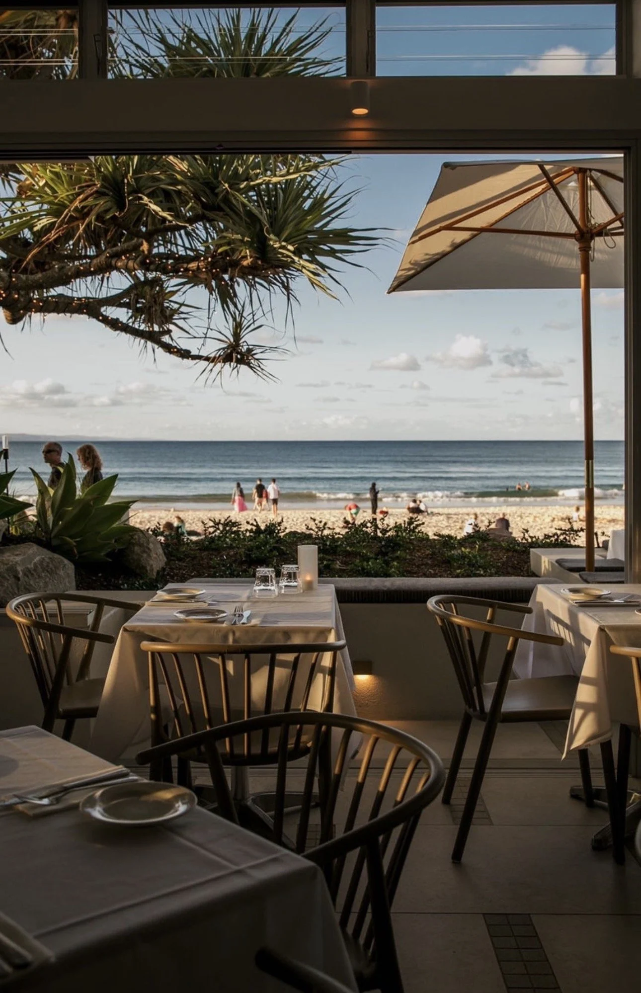 Table set for dining with plates, glasses, and utensils, overlooking a beach with people, palm trees, and an umbrella outside a restaurant.