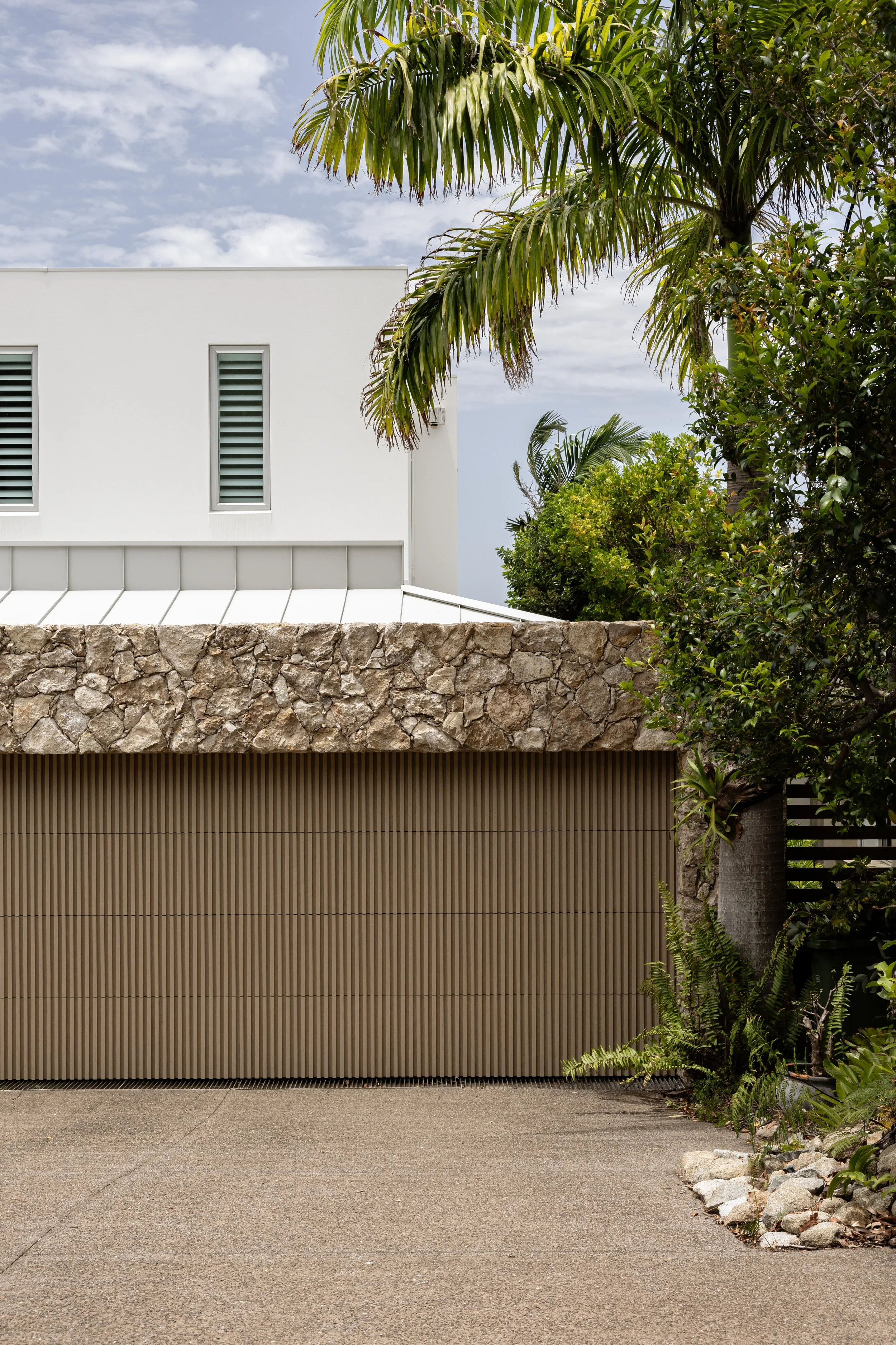 Modern house with white walls, small windows, a stone wall, beige vertical siding, and lush green palm trees and plants in the foreground, under a partly cloudy sky.