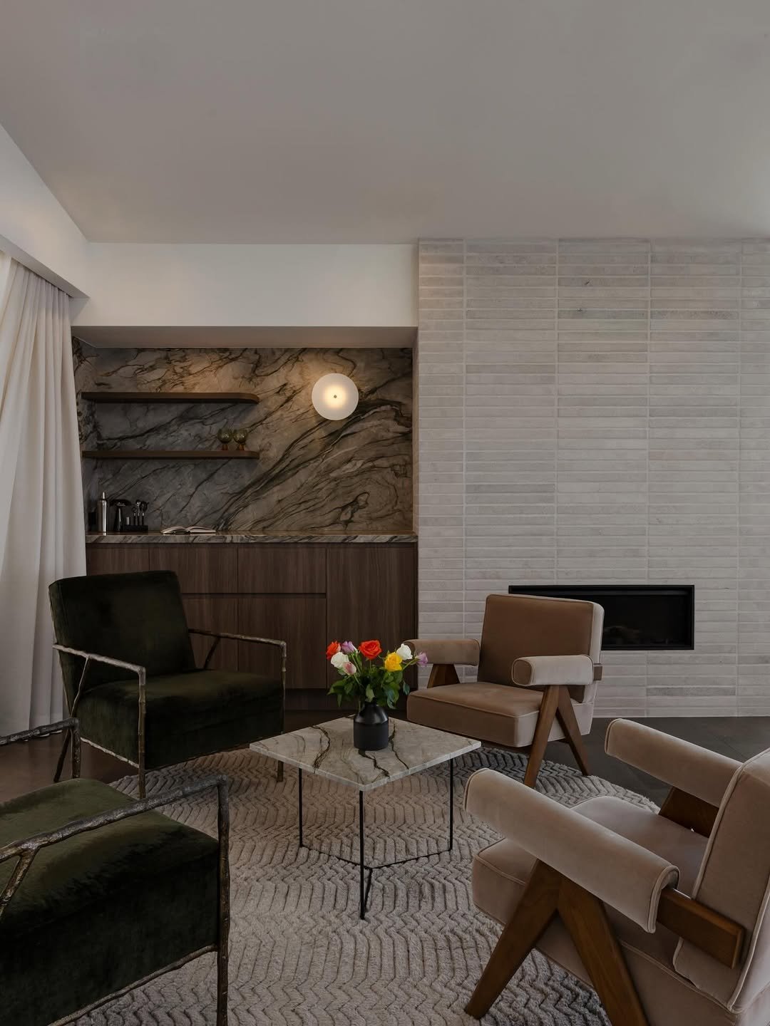 Living room with beige and dark green chairs, a marble coffee table with a vase of colorful flowers, a fireplace, and a wall with a built-in cabinet and shelves with a marble backsplash.