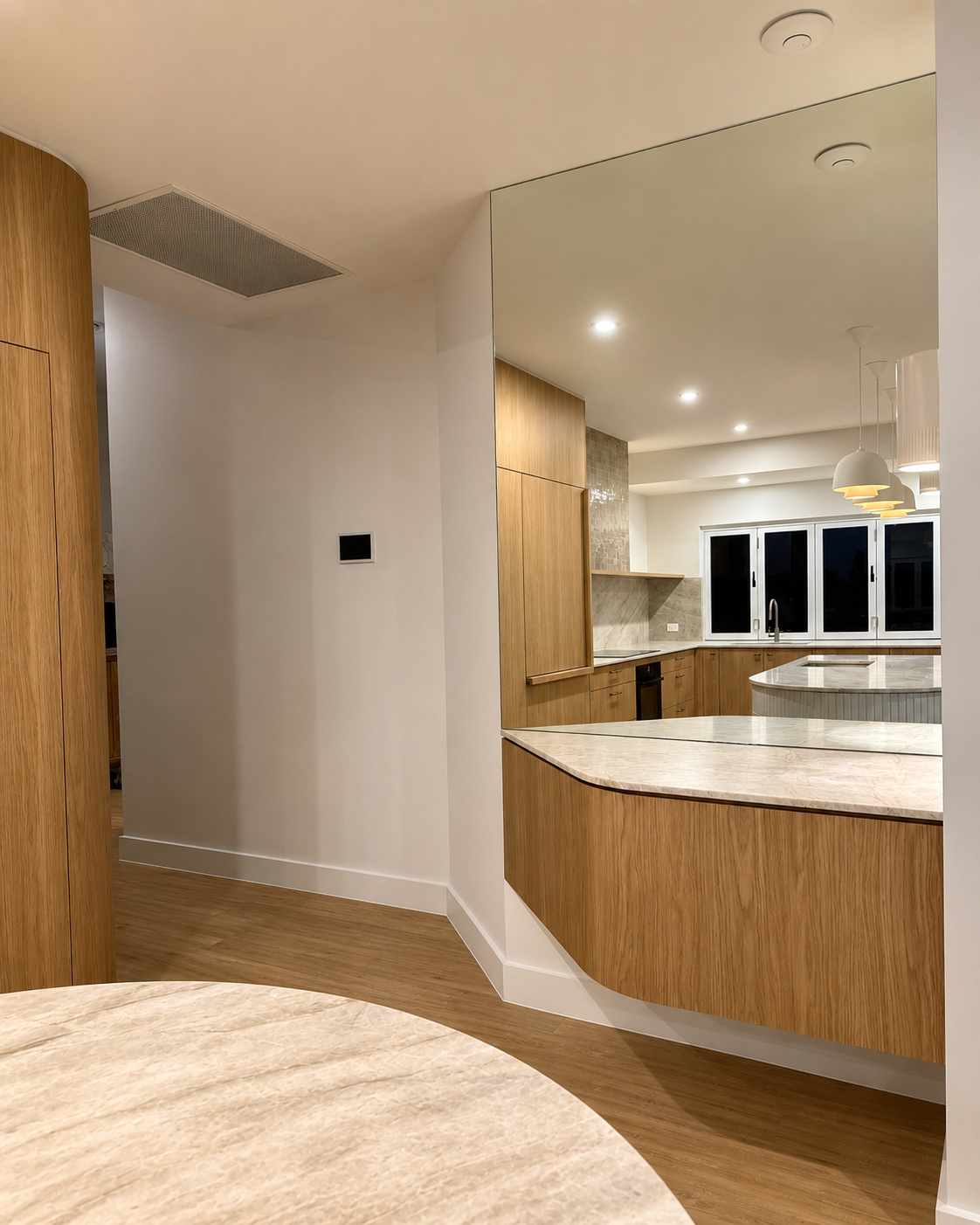 Modern kitchen with wooden cabinetry, marble countertops, and large windows at night.