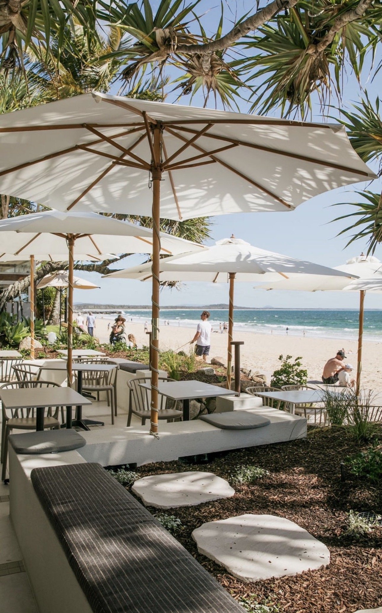 Beachside restaurant with white tables, chairs, and large white umbrellas, overlooking the sandy beach and ocean, with people walking and relaxing near the shoreline under a clear blue sky.