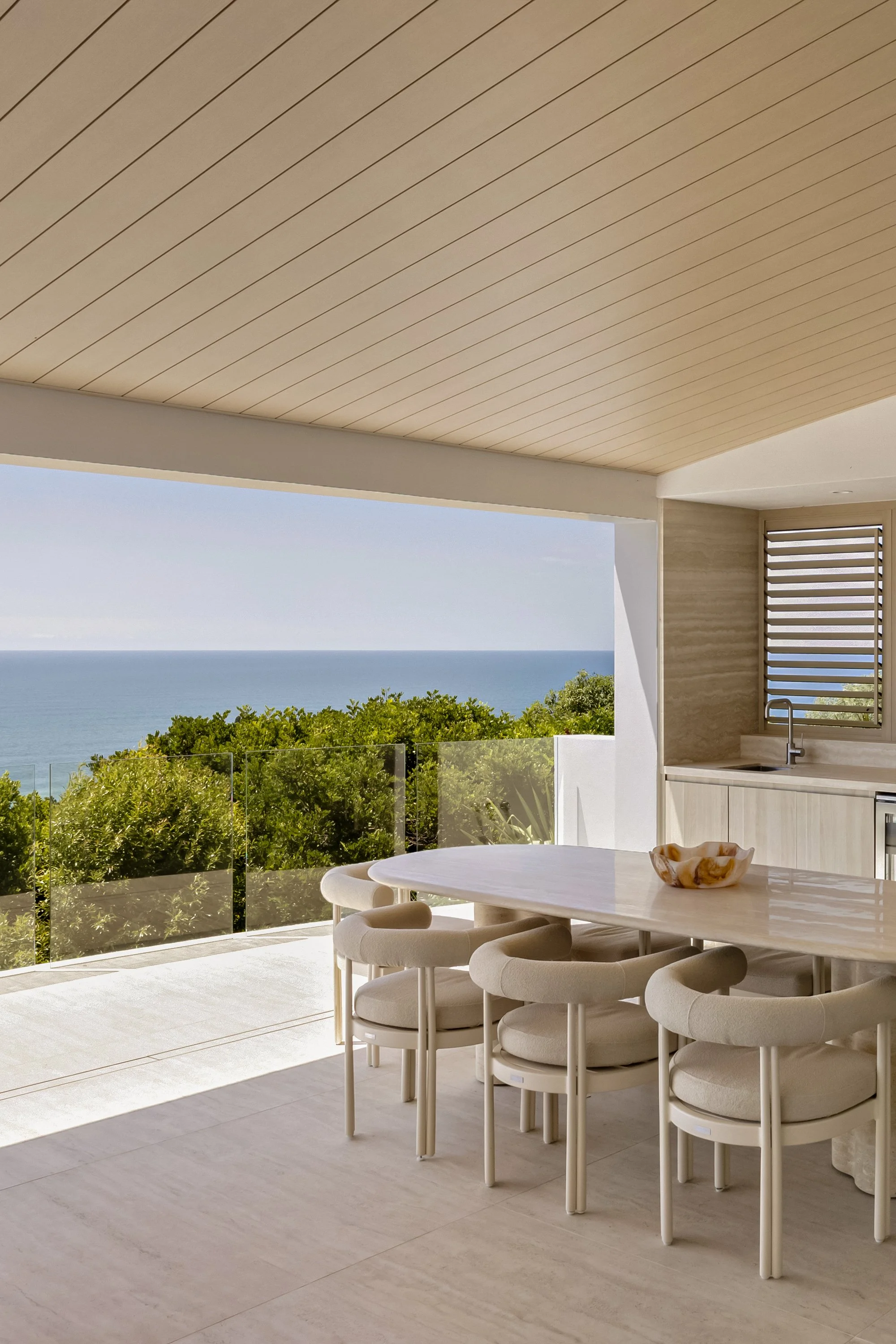 Modern outdoor dining area with a large white marble table, cushioned chairs, and a view of the ocean and green trees in the distance, under a beige ceiling with a slatted design.