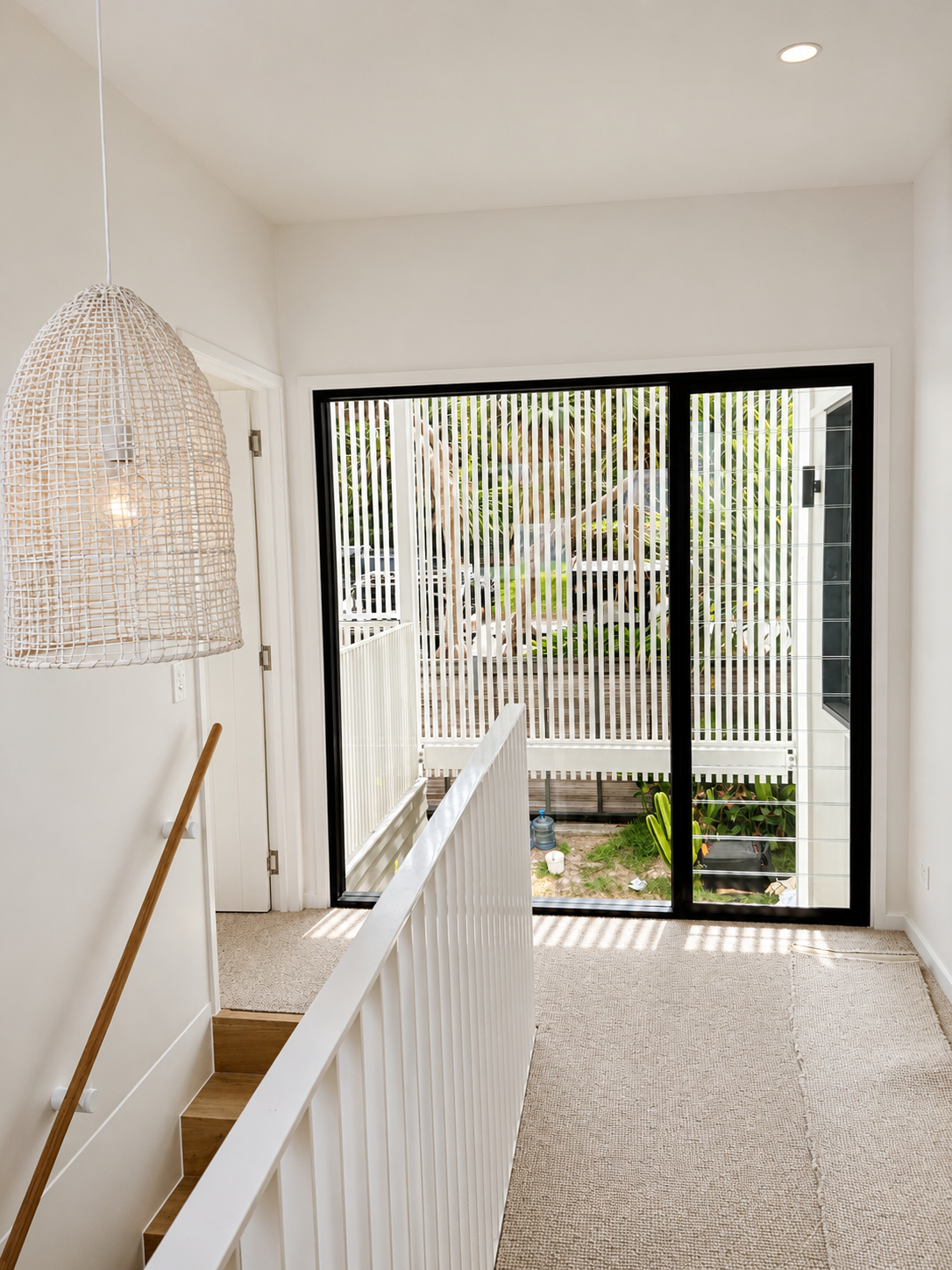 Interior view of a stairway landing with a white railing, beige carpet, a woven hanging lamp, and a large sliding glass door with vertical blinds leading outside to a small yard.