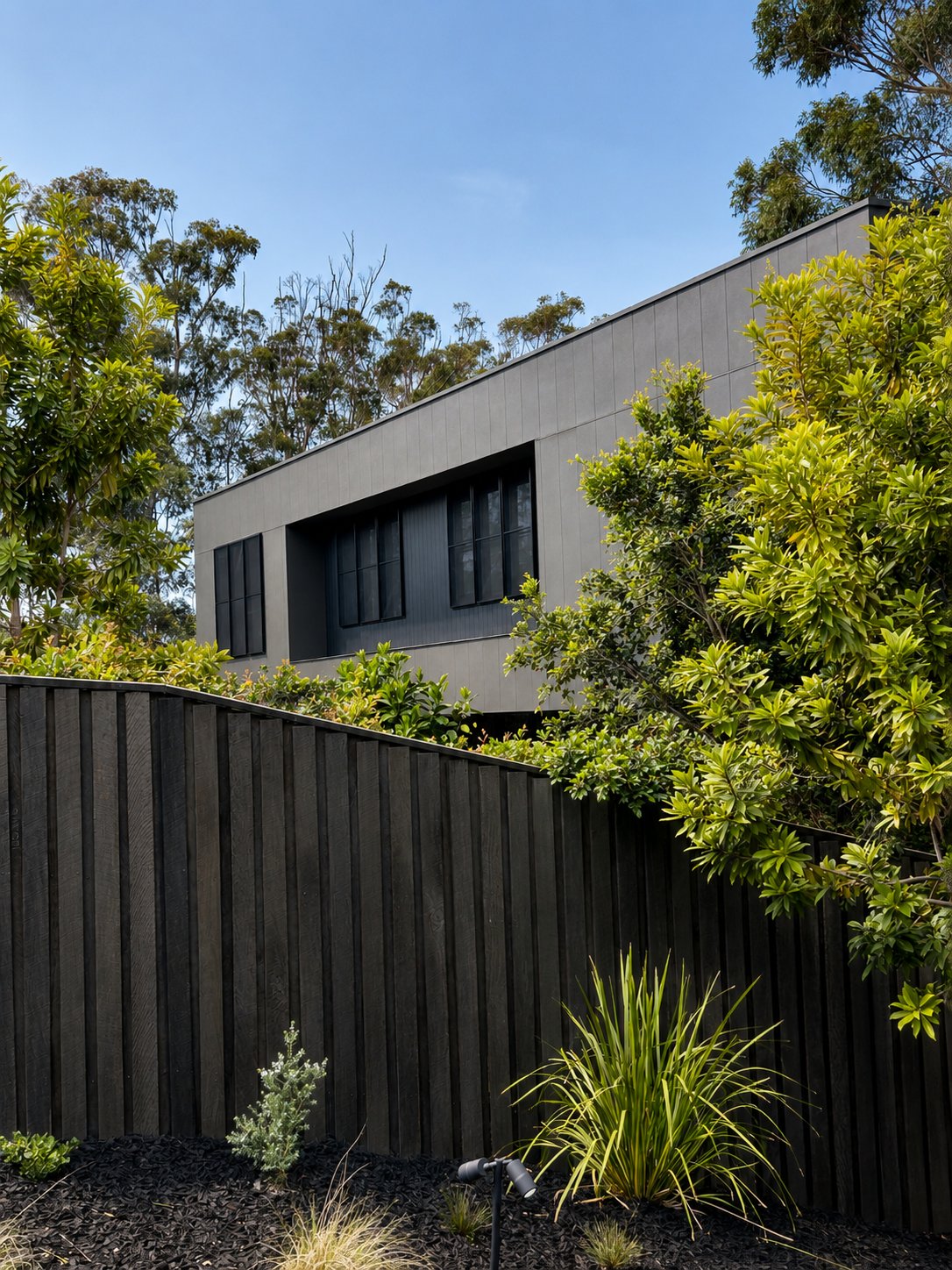 Modern house with large black windows, gray exterior, surrounded by lush green trees and shrubs, with a wooden fence and landscaped garden in the foreground.