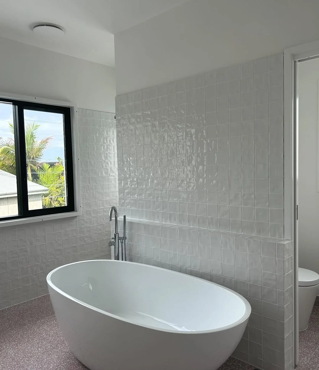 Modern bathroom with a freestanding bathtub, window showing palm trees outside, and a toilet in an adjacent room.