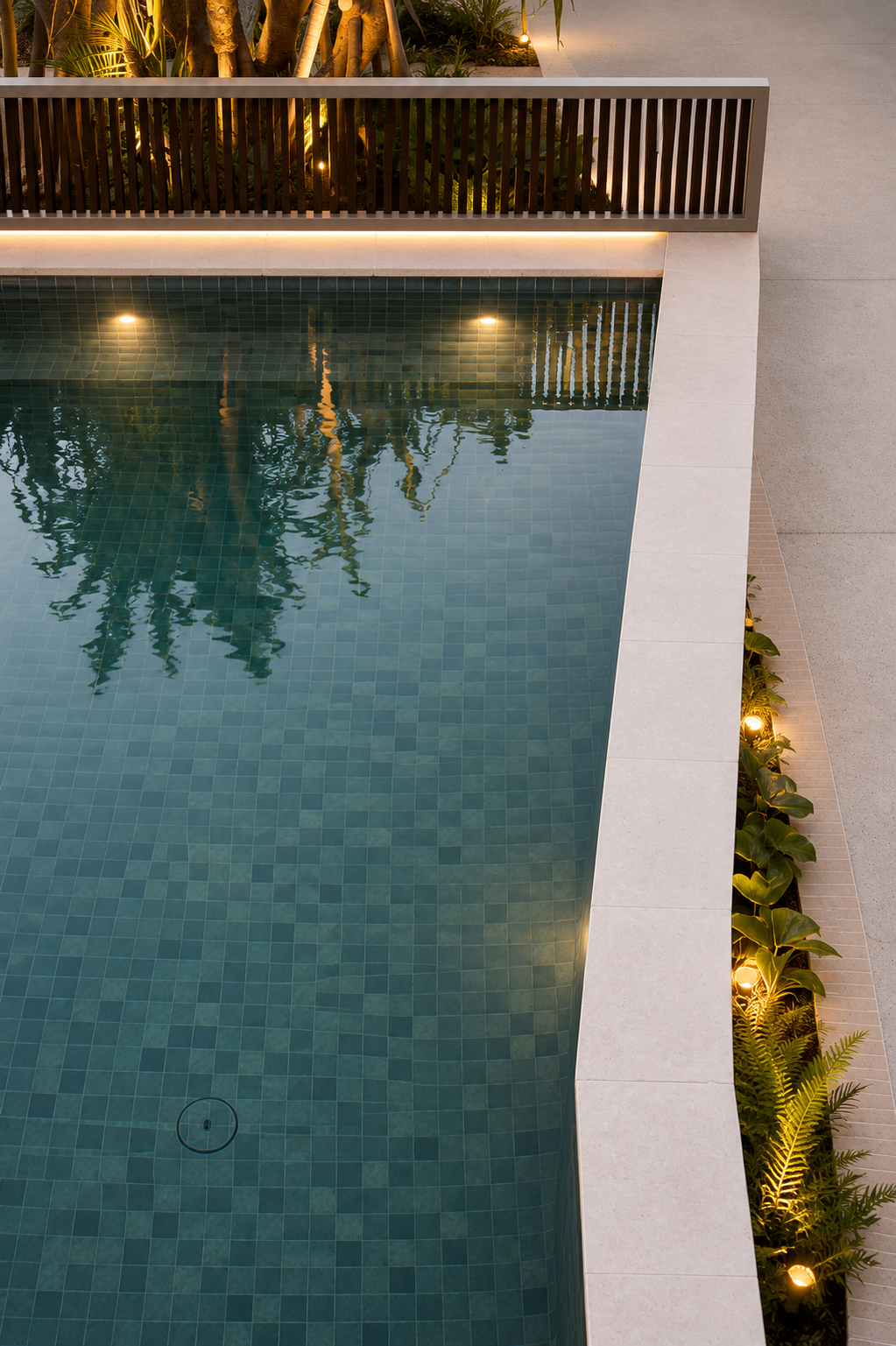 A modern swimming pool with blue tiles, surrounded by a white concrete border, with plants and outdoor lighting along the edge.