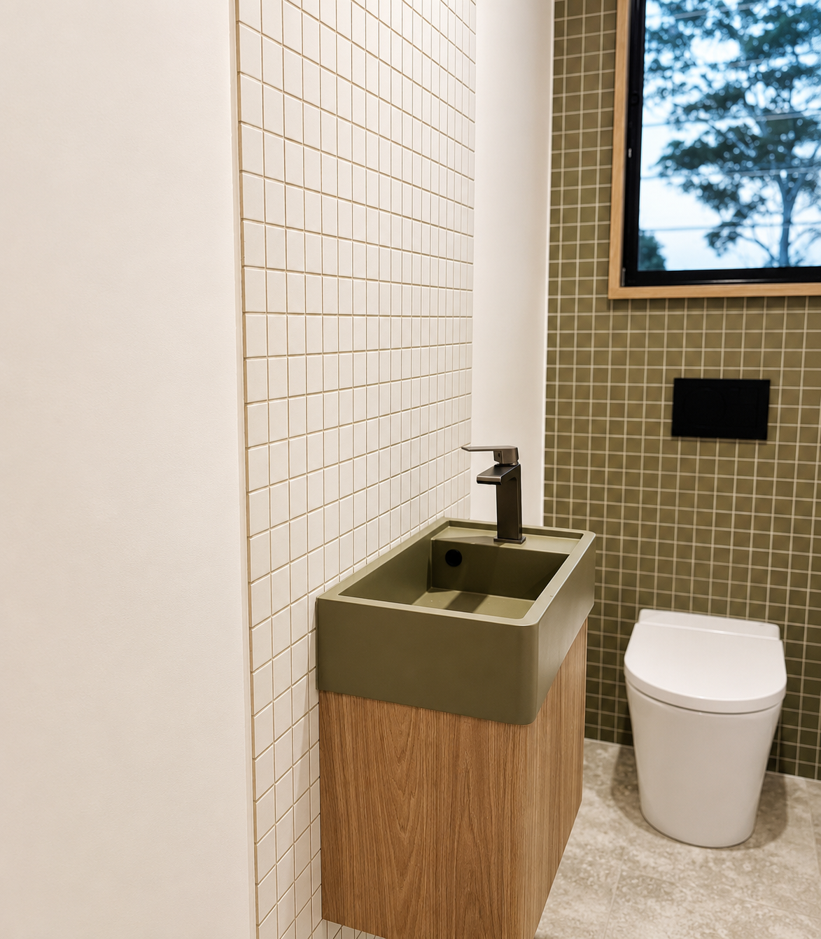 Small bathroom with a green sink, black faucet, and a toilet, with beige and green tiled walls and a window showing trees outside.