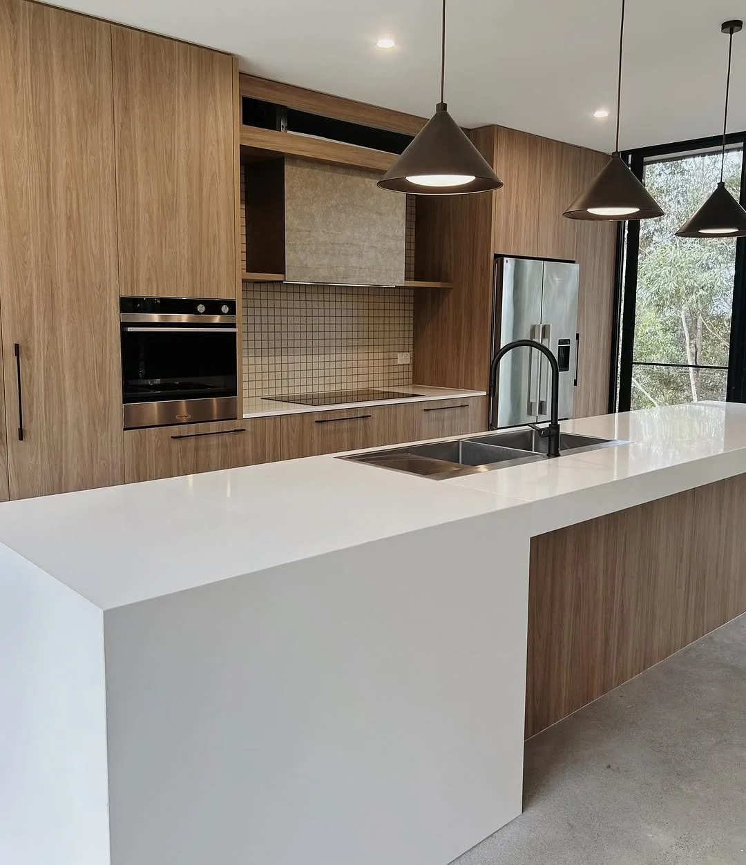Modern kitchen with wood cabinetry, white island, stainless steel refrigerator, oven, and black pendant lights.