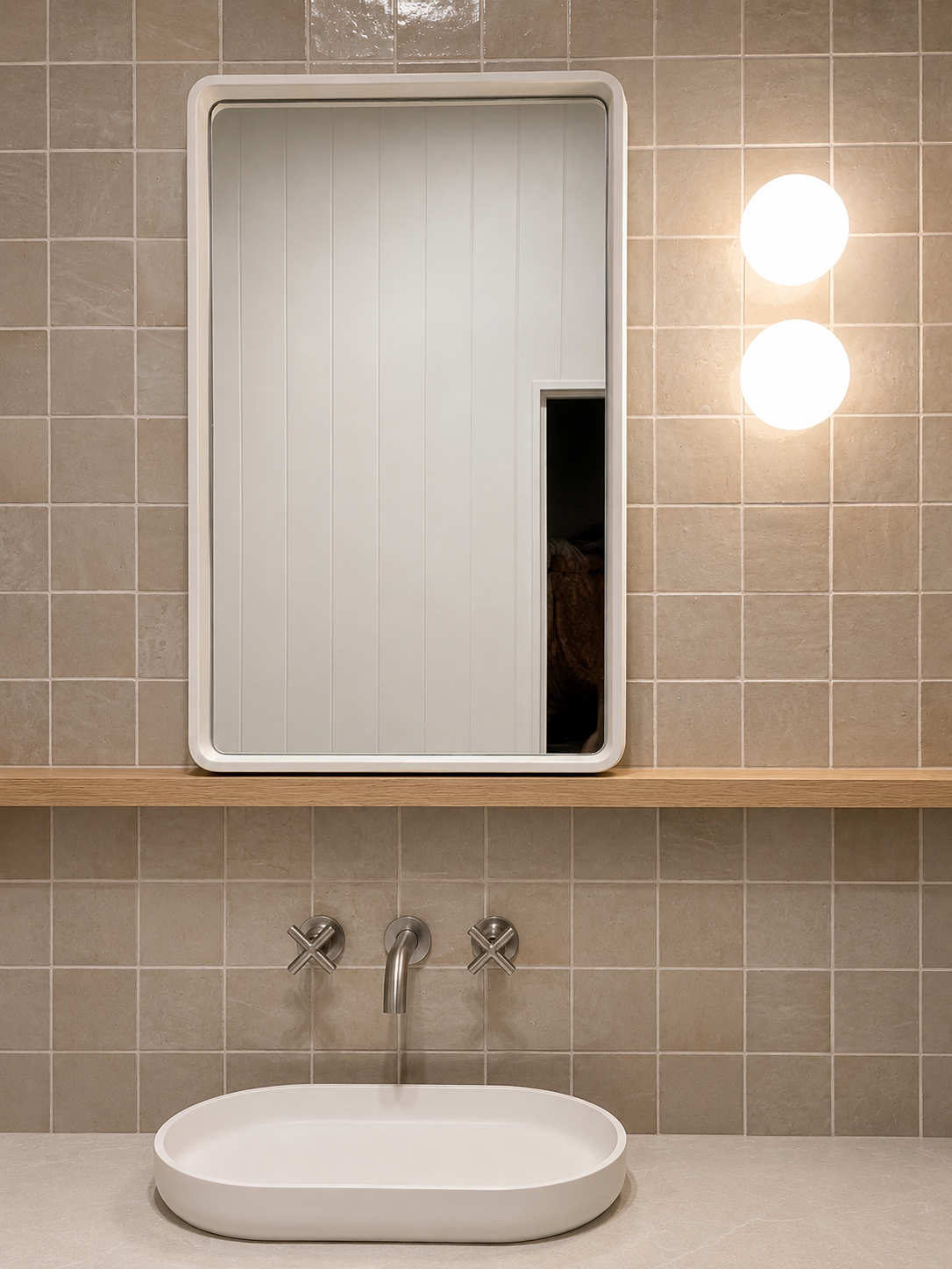 Modern bathroom with a white oval sink, a wall-mounted mirror, and two round wall lamps on a tiled beige wall.