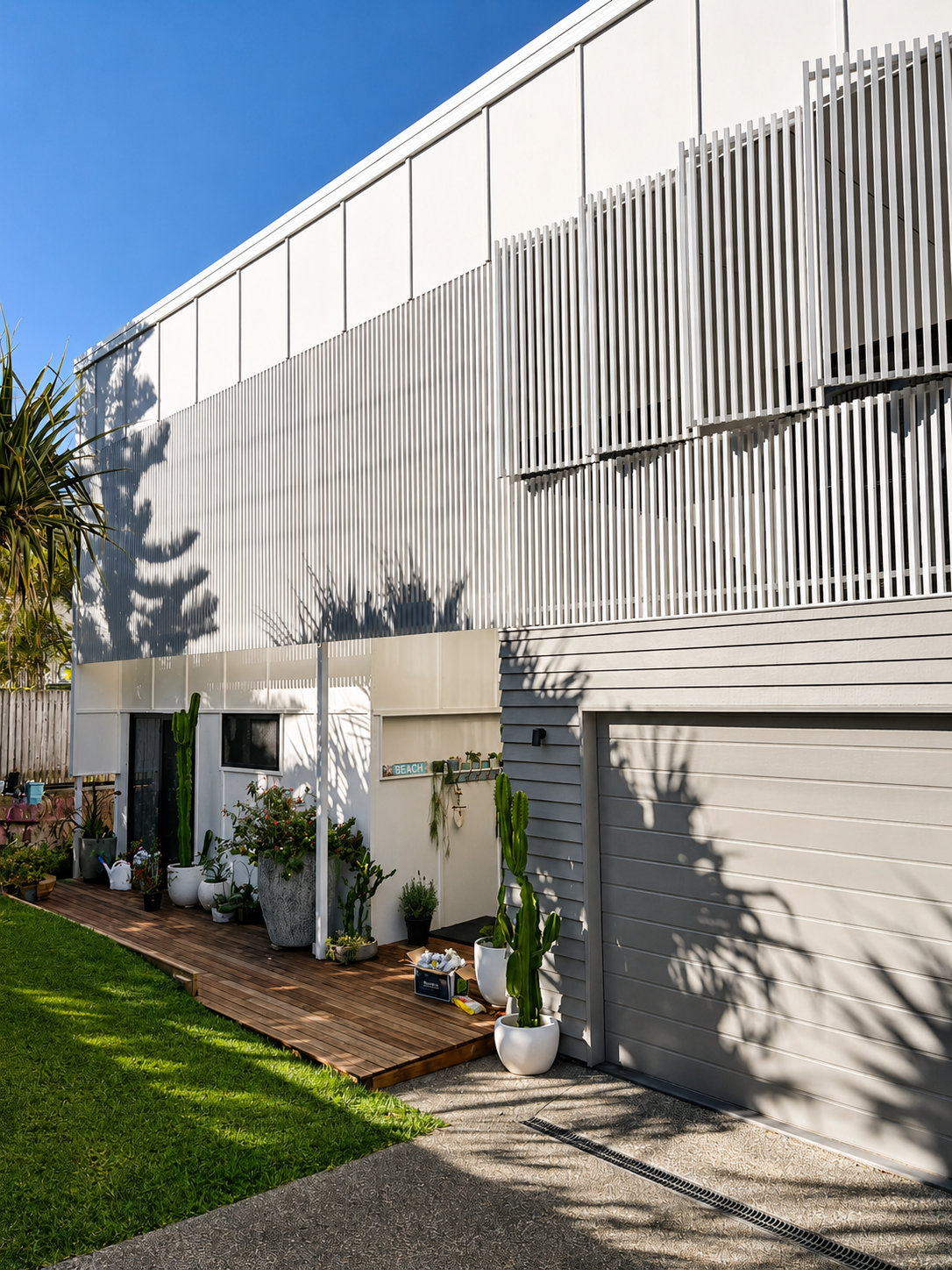 Modern white house with horizontal siding and tall, narrow white slatted panels, small deck with potted cactus plants, and green lawn.