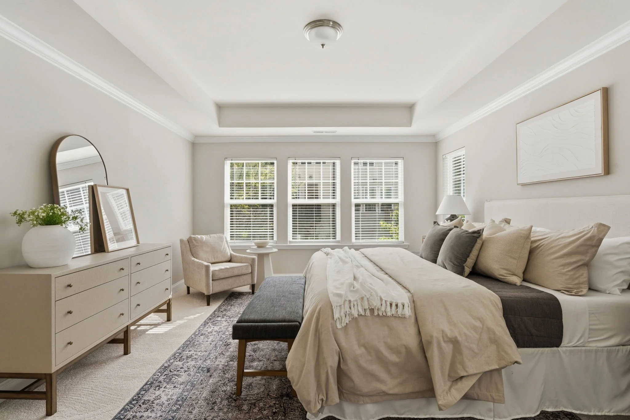 Bedroom with off-white walls, beige carpeted floor, a white bed with neutral pillows and comforters, two wooden chairs, a side table with lamp, and a a window and a dresser. .