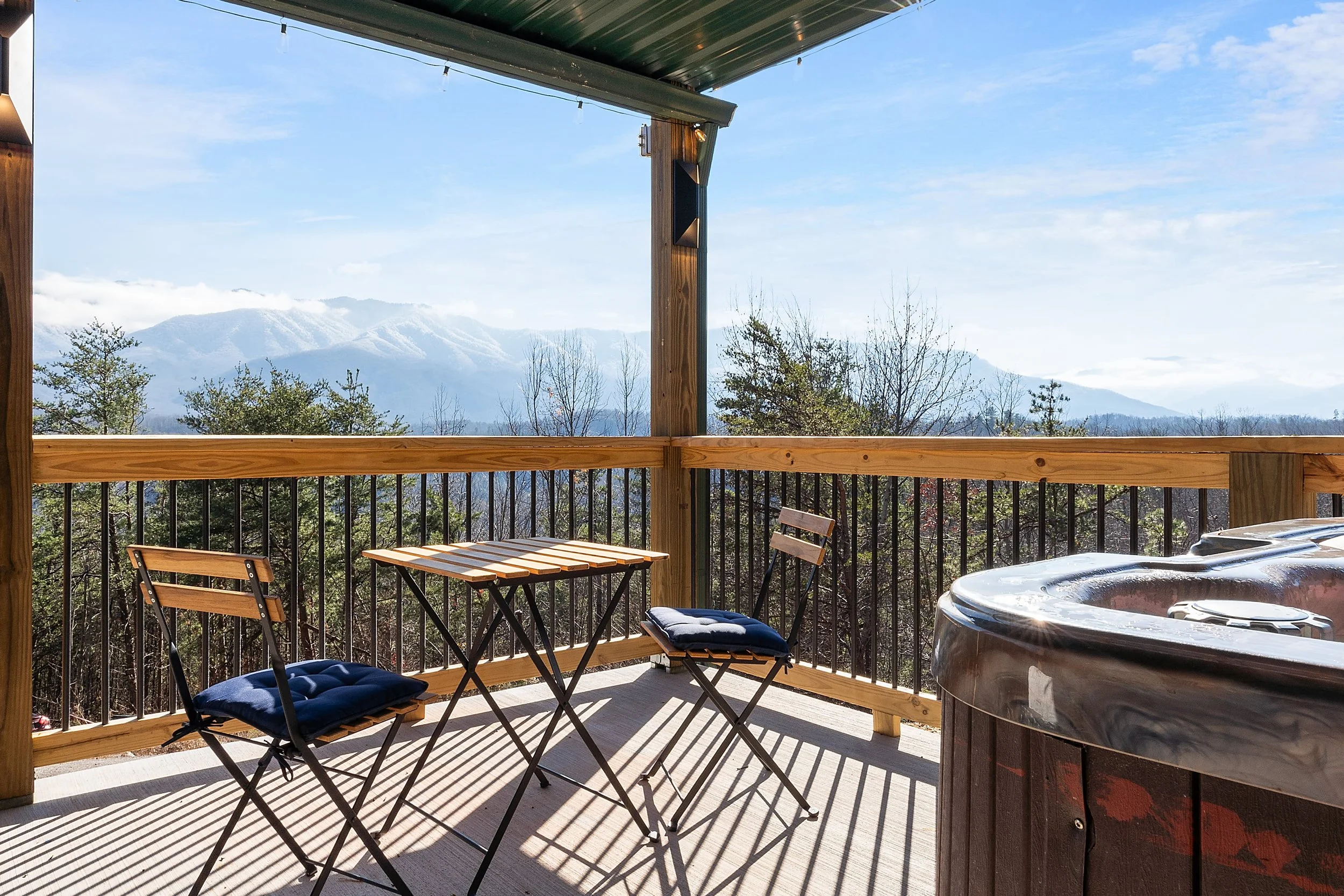 A balcony with a small wooden table and four folding chairs with navy cushions, overlooking trees and snow-capped mountains in the distance on a sunny day