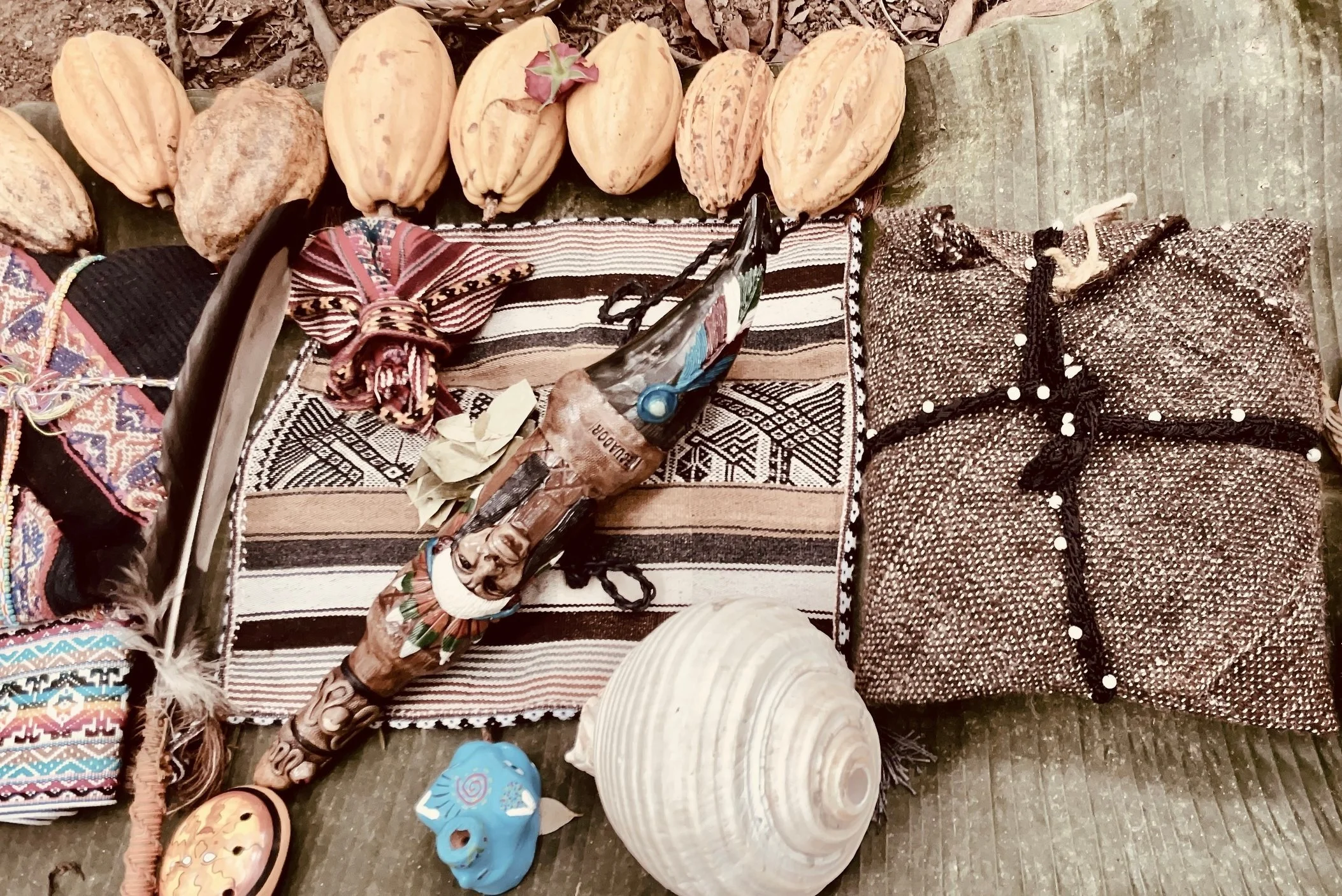 Mesa de objetos rituales con cacao, textiles tradicionales, plumas, hojas de coca, cetro decorativo y concha de caracol.