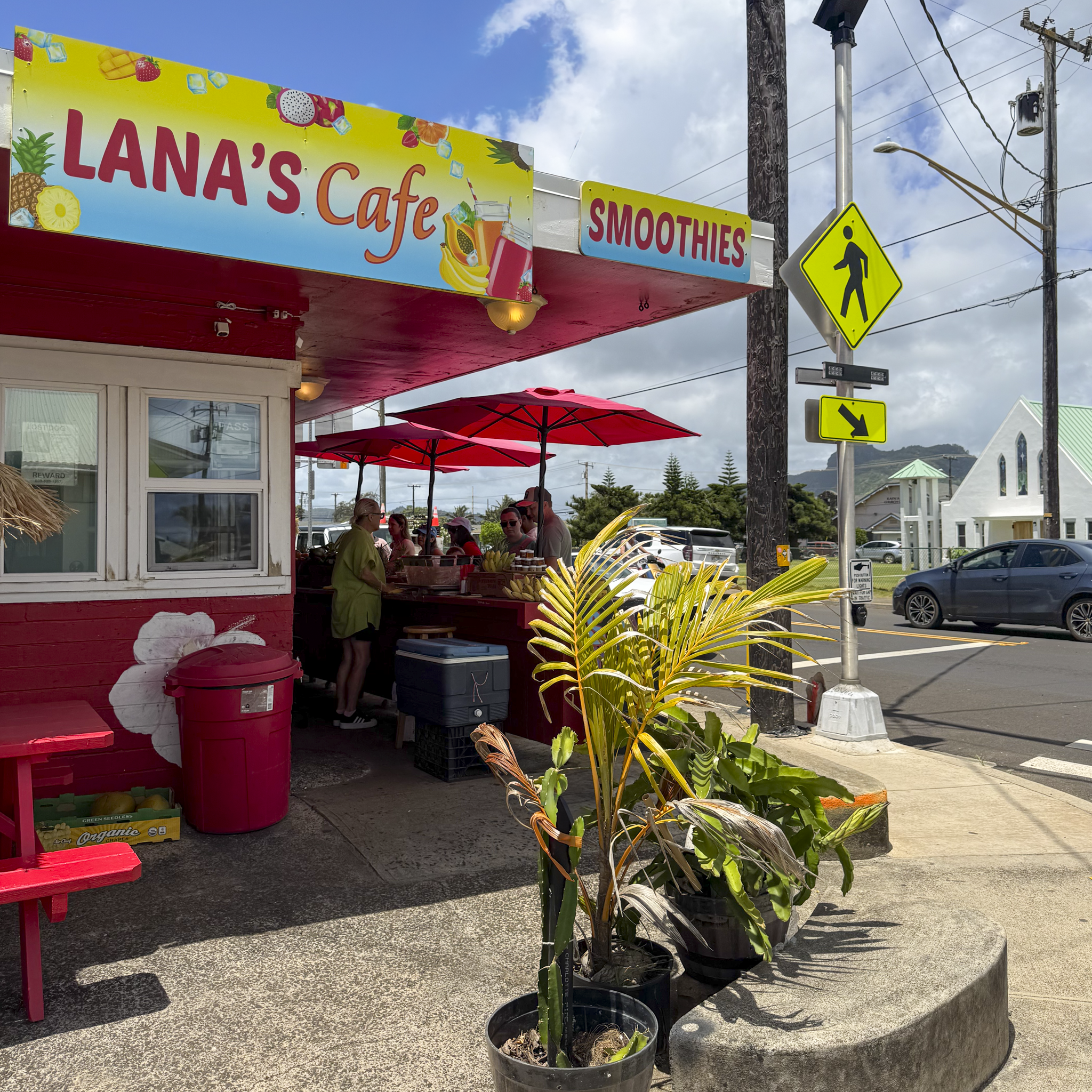 Bright yellow and pink sign for 'Lana's Cafe' and smoothies, with images of fruits and beverages, outside a cafe with red umbrellas and customers, next to utility poles and street signs, under partly cloudy sky.