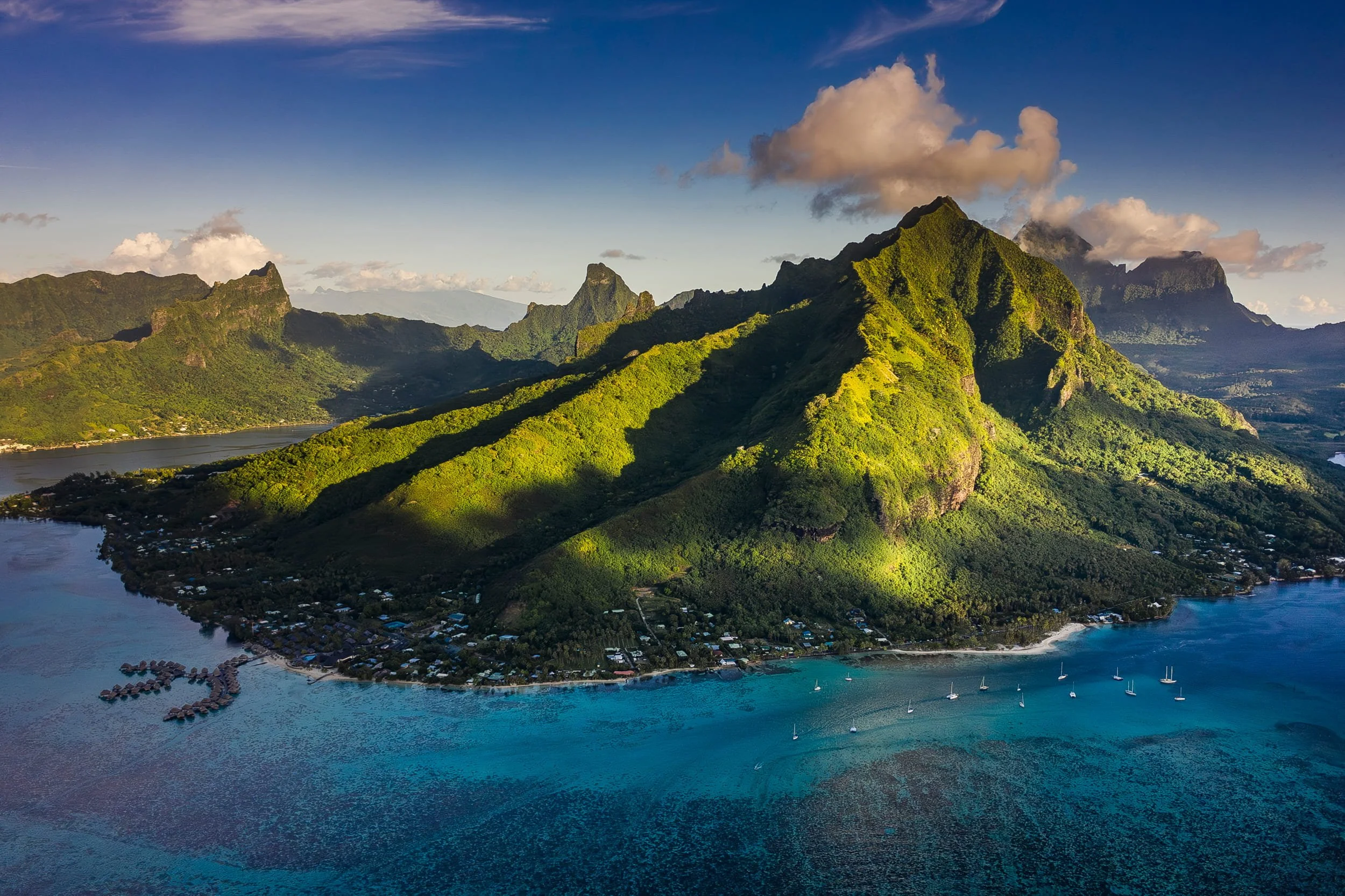 Aerial view of lush green mountains and water, with a small overwater bungalow resort and boats near the shoreline.