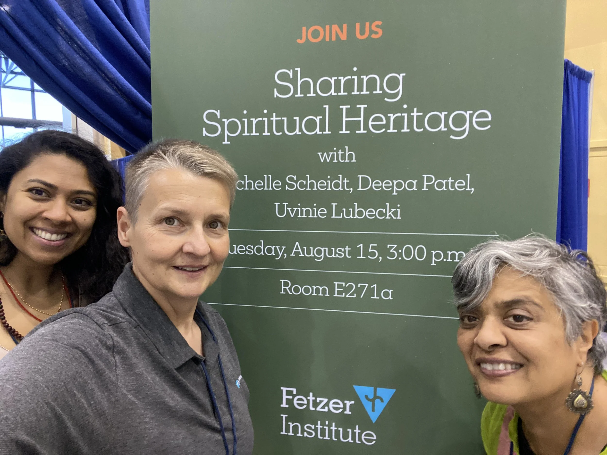 Three people smiling in front of a sign for an event titled "Sharing Spiritual Heritage" hosted by the Fetzer Institute, scheduled for Tuesday, August 15, 3:00 p.m., Room E271a.