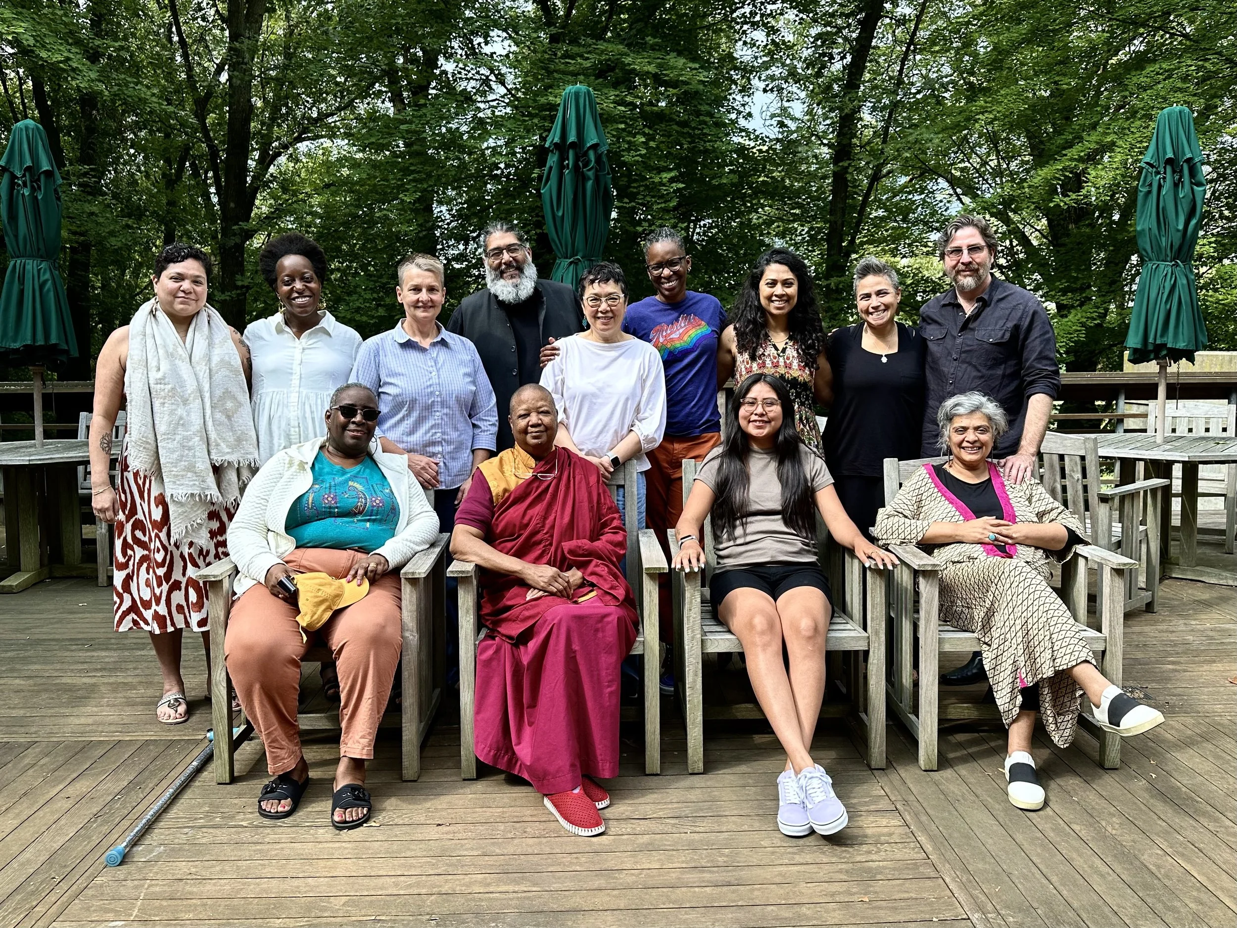 Group of diverse people sitting and standing on a wooden deck with green umbrellas, surrounded by trees.