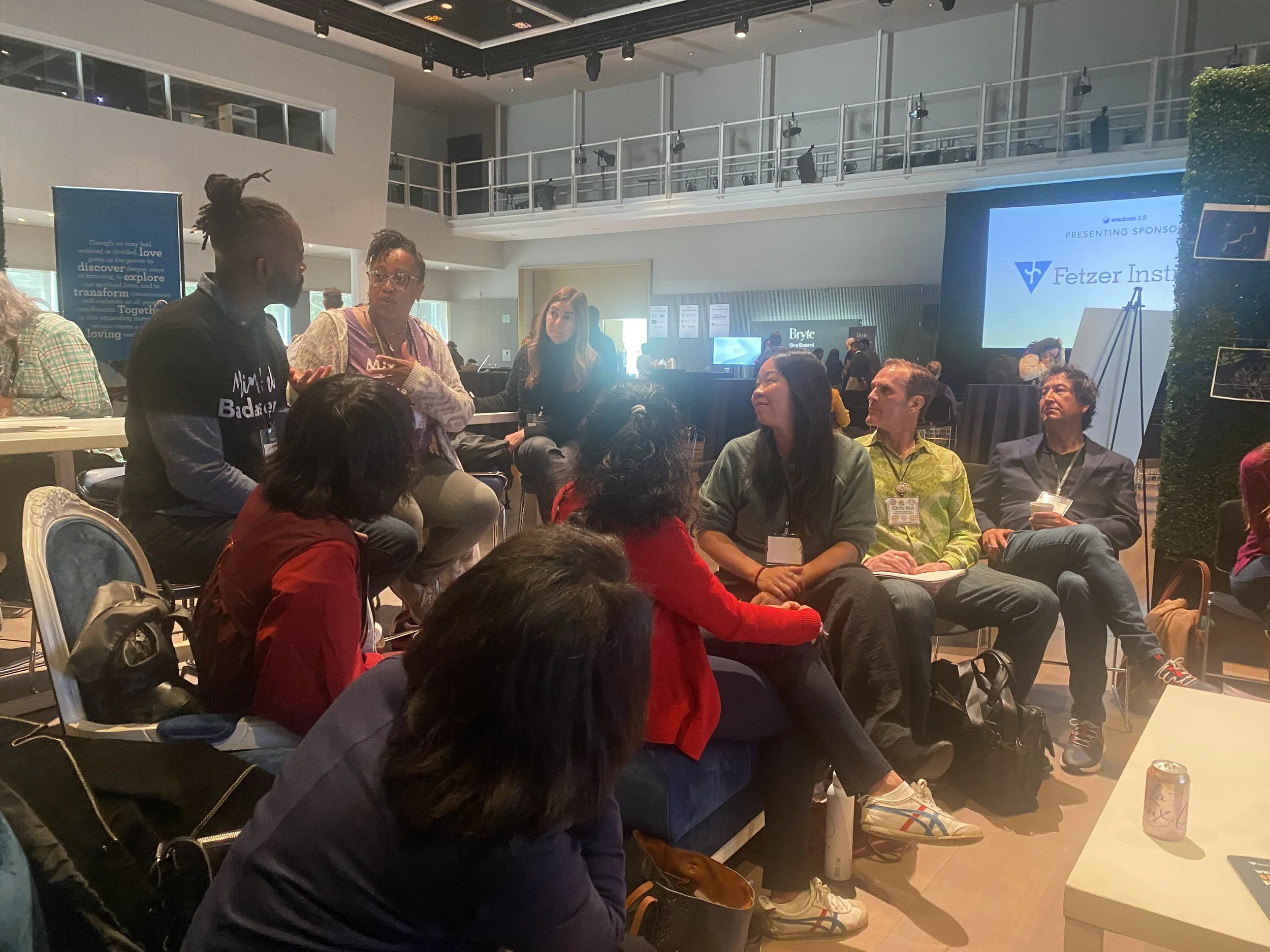 Group discussion at a conference, with diverse people seated in a circle in a modern venue. A screen in the background displays "Fetzer Institute."