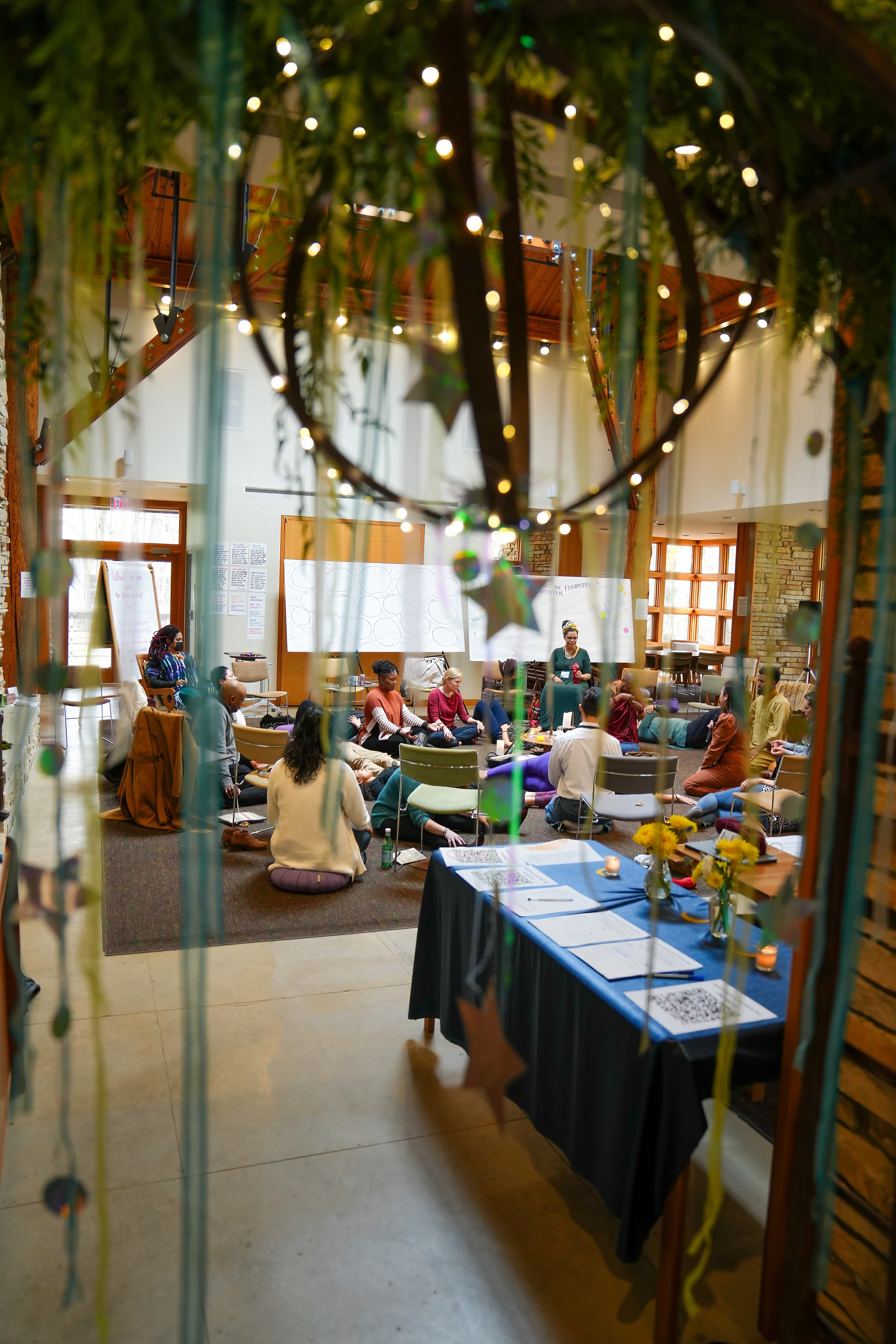 People sitting in a circle in a room with plants and lights overhead, focusing on a group activity or discussion.