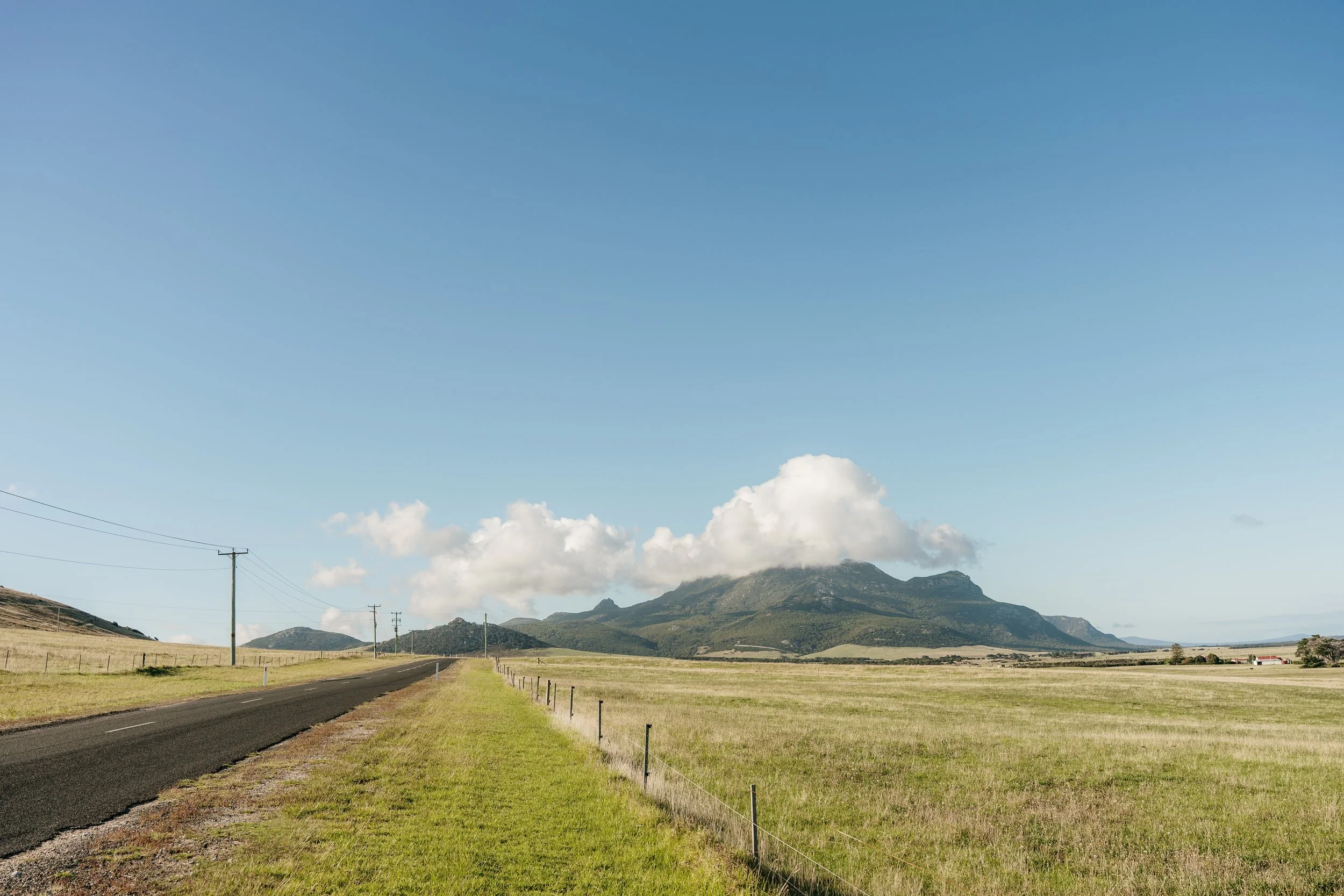 A rural landscape with a paved road on the left, green grass fields separated by fencing, rolling hills, and mountains in the background under a blue sky with some clouds.
