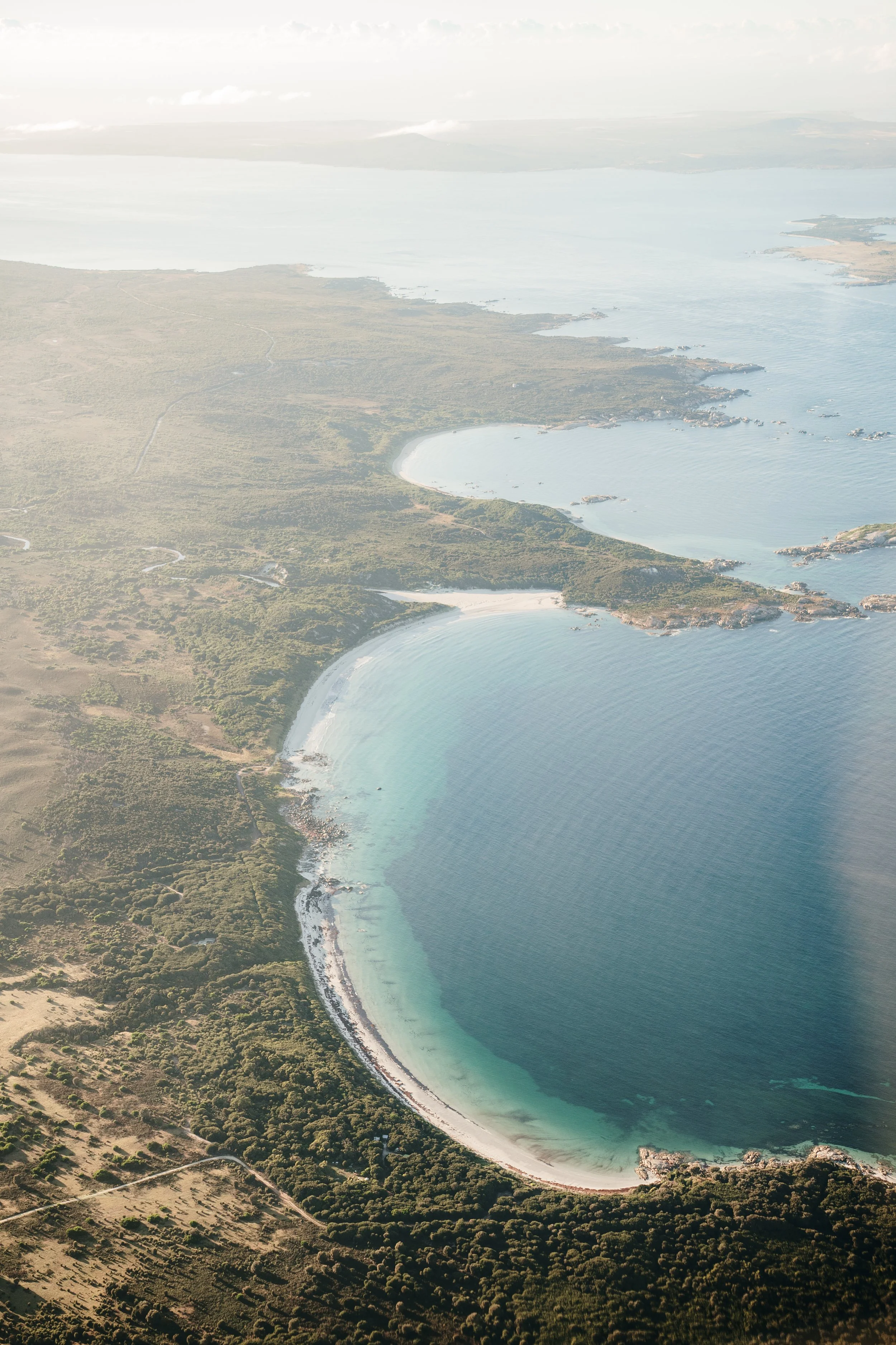Aerial view of a curved sandy beach along a turquoise lagoon with dense green vegetation and a few small buildings, overlooking a large body of water extending to distant landmasses under a cloudy sky.