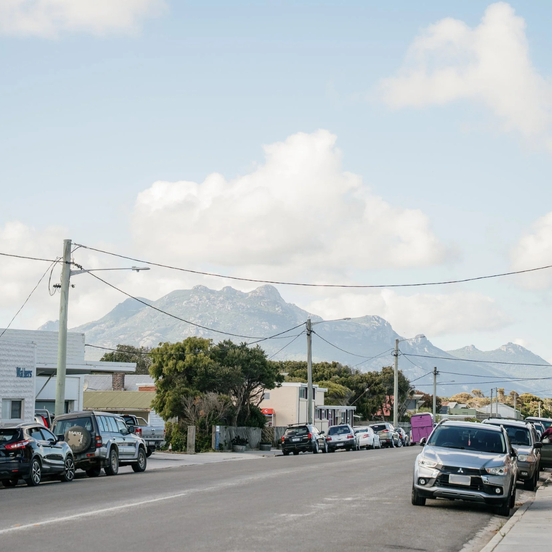 Street view with parked cars, power lines, buildings, trees, and mountains in the background under a partly cloudy sky.