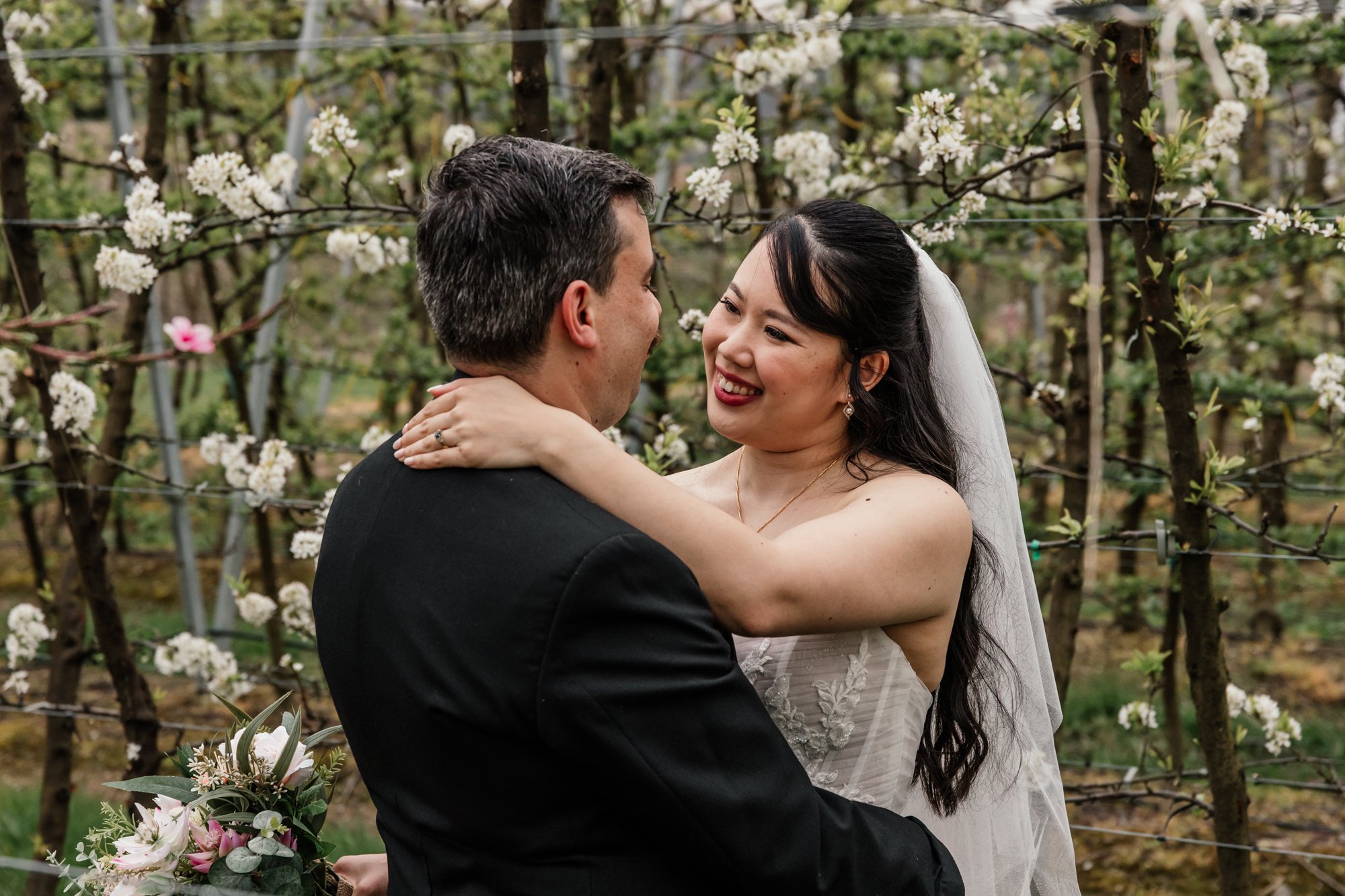 A bride and groom embrace in a garden or orchard with blooming flowers, smiling at each other.