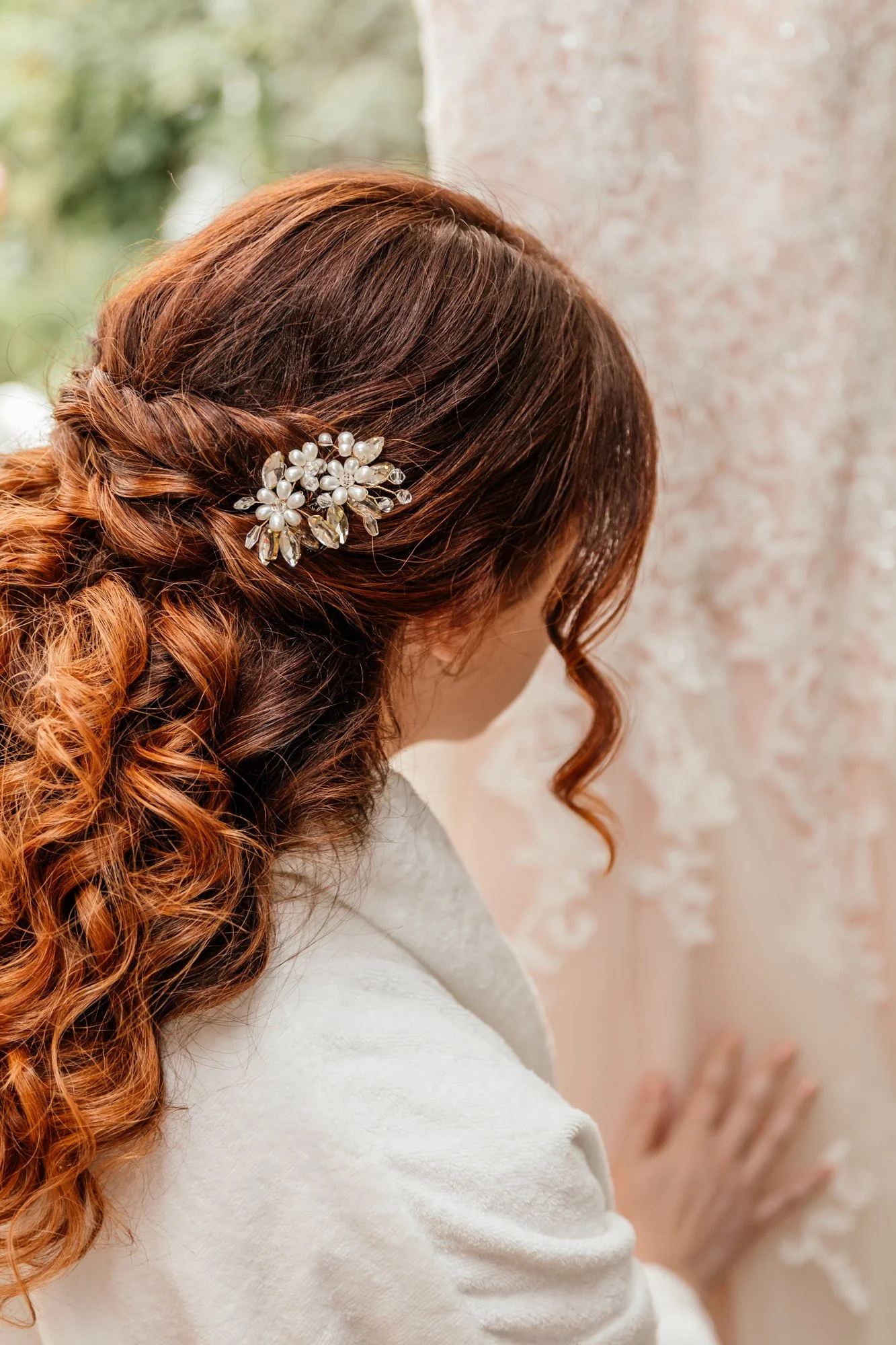 A woman with long, curly, red hair styled with a pearl and crystal floral hair clip, wearing a white coat, against a soft pink floral background.