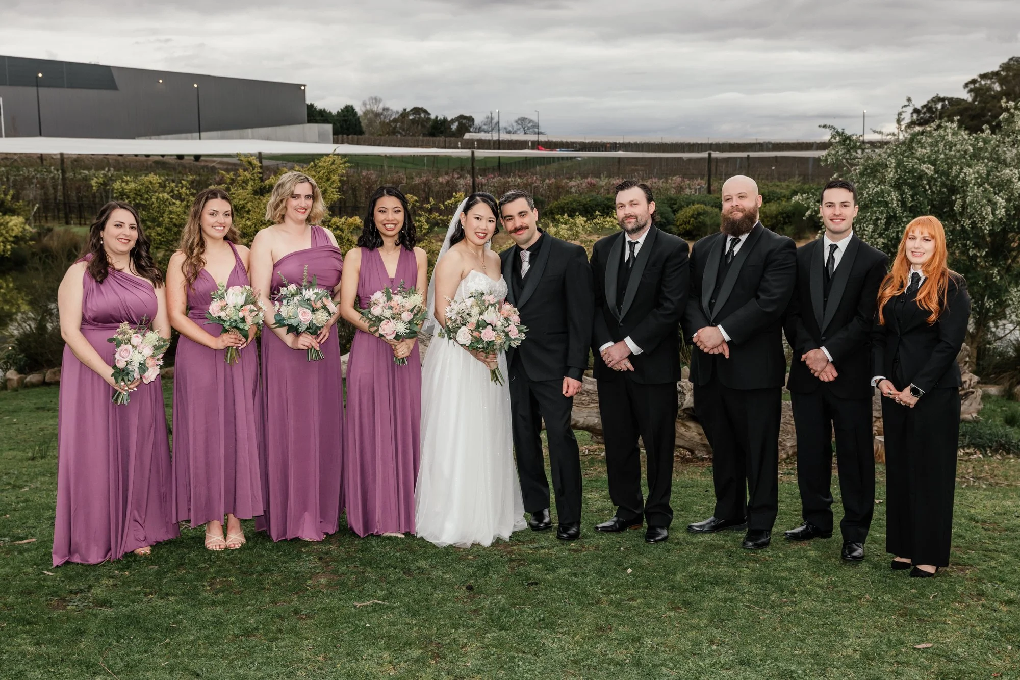 A group of nine people at a wedding outdoors, including bridesmaids in purple dresses, a bride in a white gown, and groomsmen in black suits, standing on a grassy area with trees and a cloudy sky in the background.
