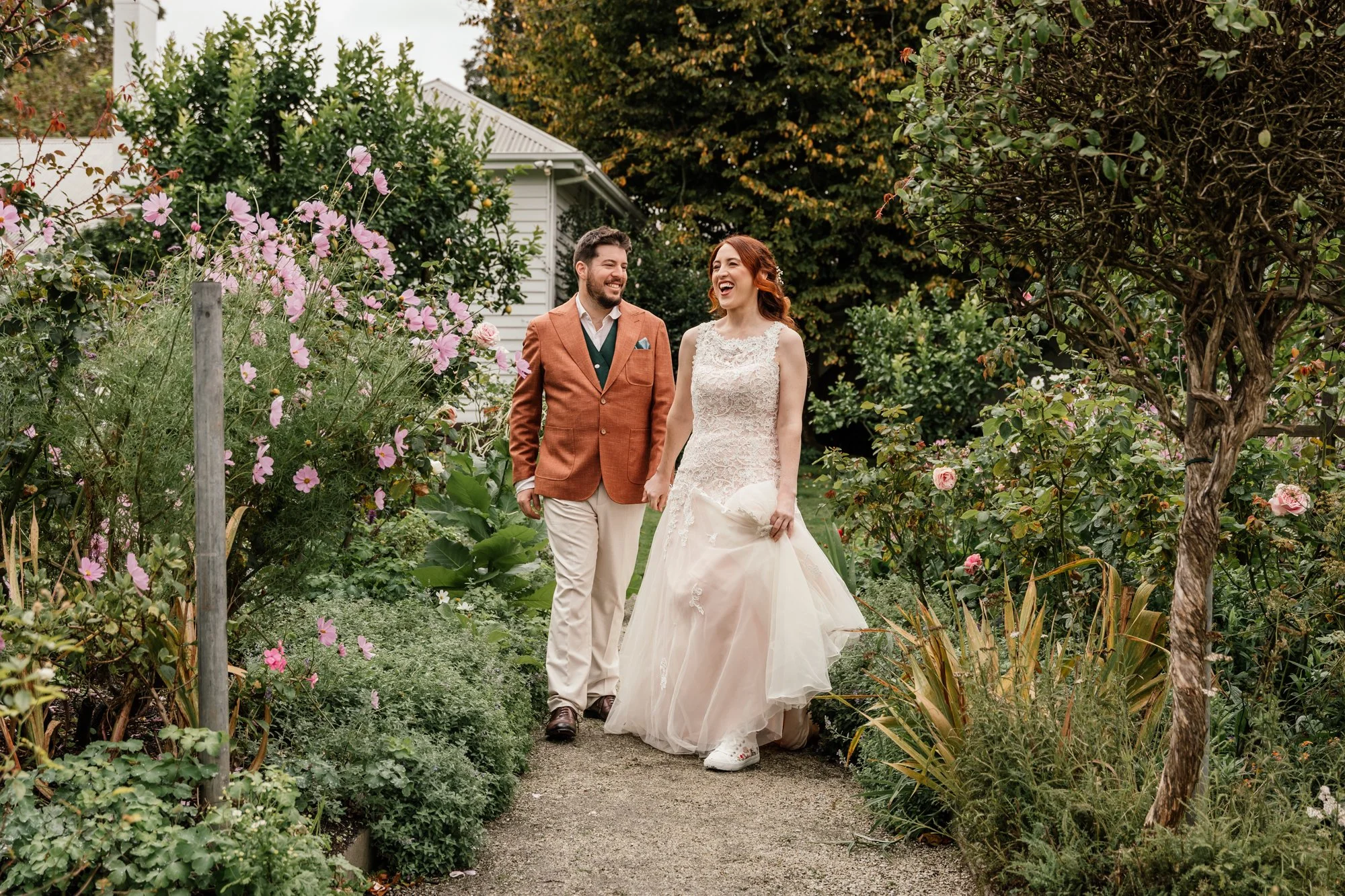 A bride and groom walking through a garden with lush green plants and blooming flowers, holding hands and sharing a joyful moment.